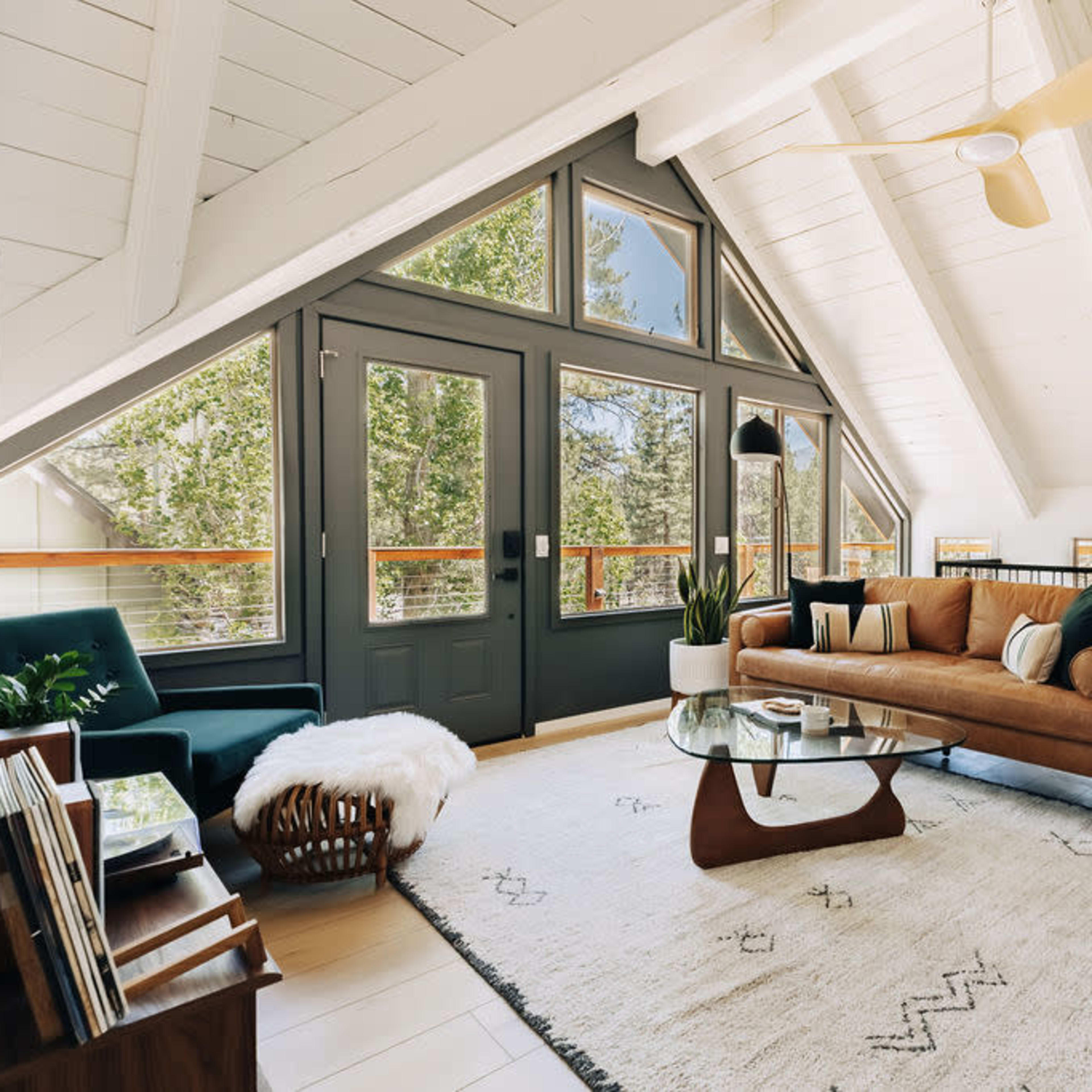 A modern living room features a brown sofa, a glass coffee table, and large windows framed in dark blue, set under a vaulted ceiling with wooden beams.