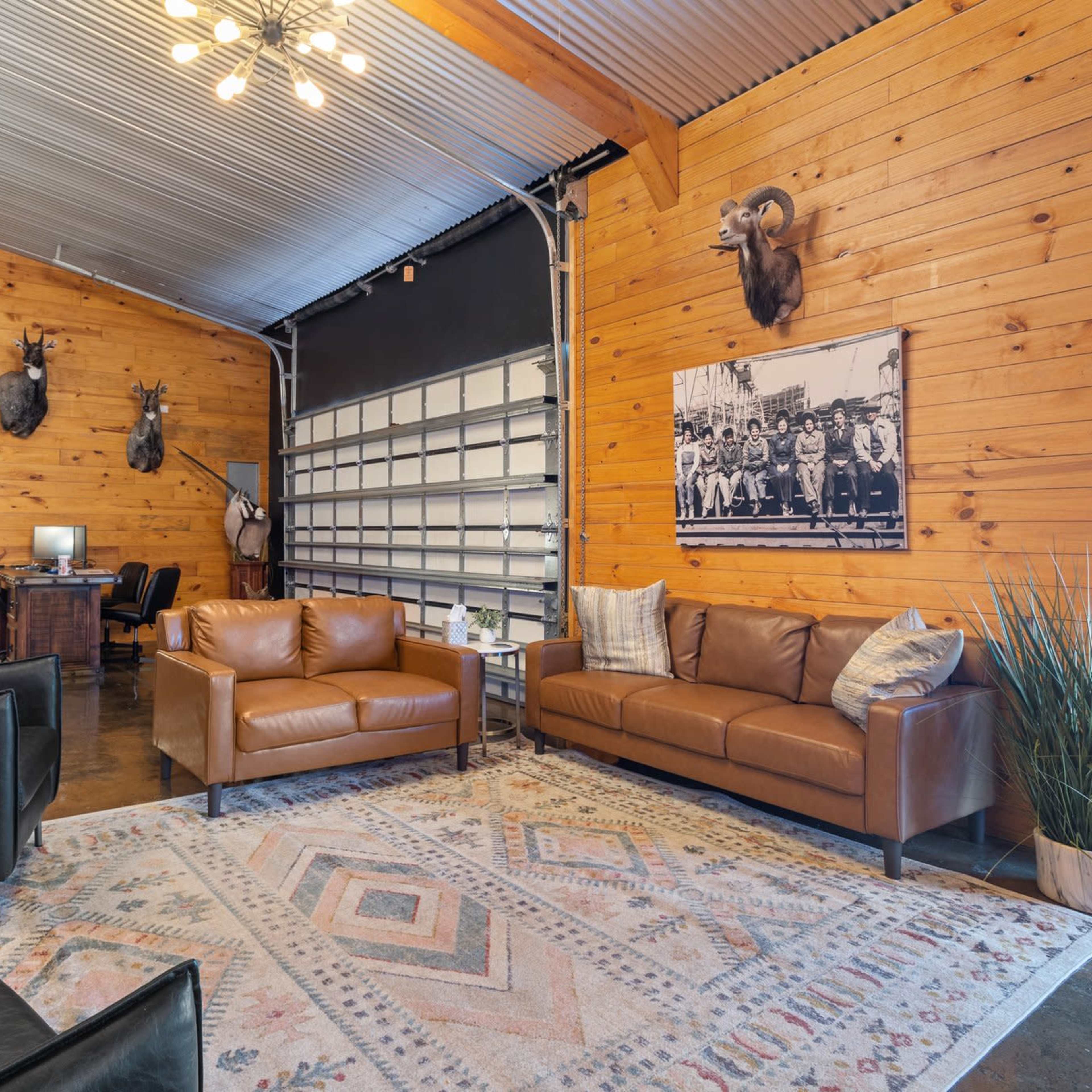 A spacious lounge with wooden walls, featuring mounted animal heads, leather sofas, a large rug, and a desk area.