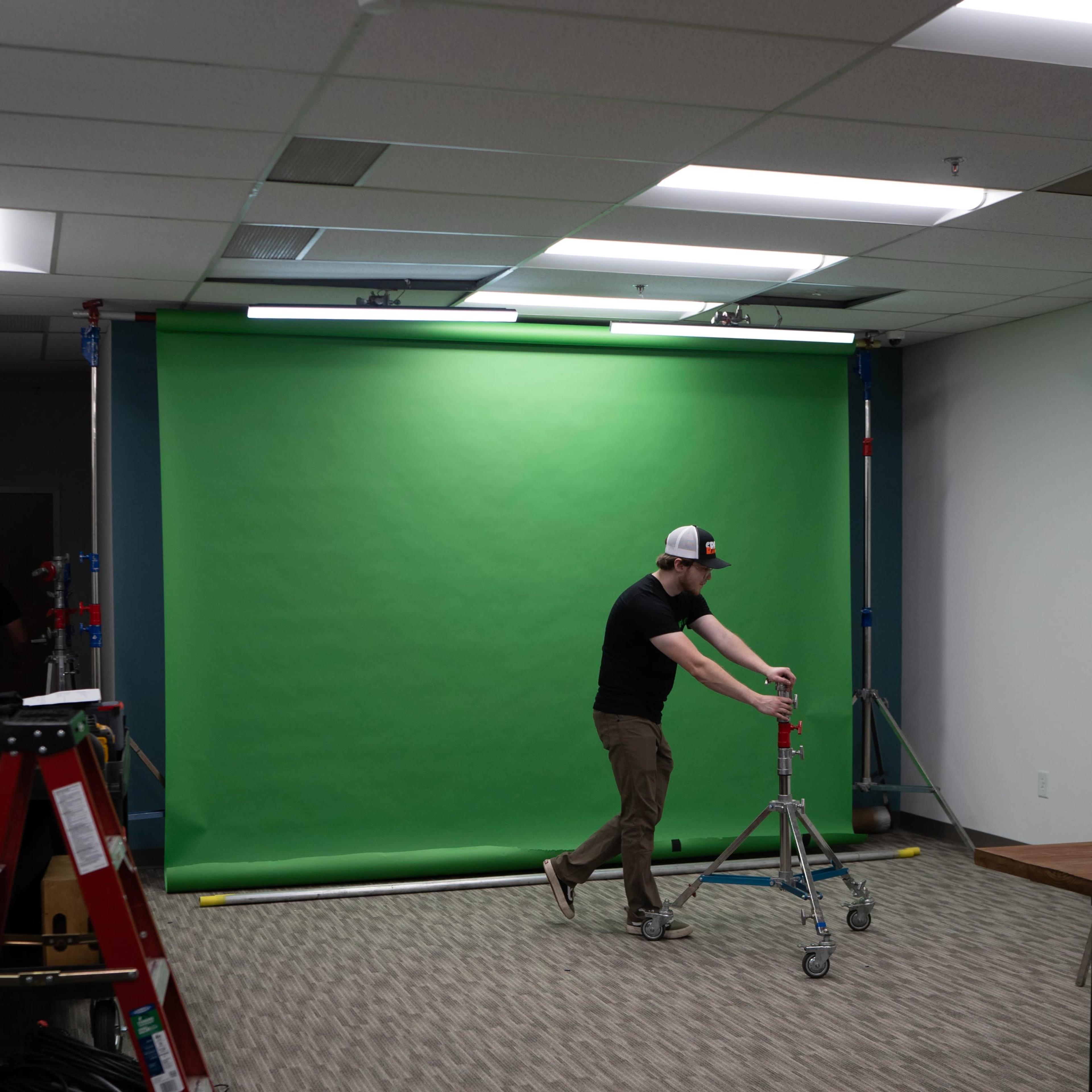 A person adjusts a tripod in front of a green backdrop in a room with carpeted flooring.