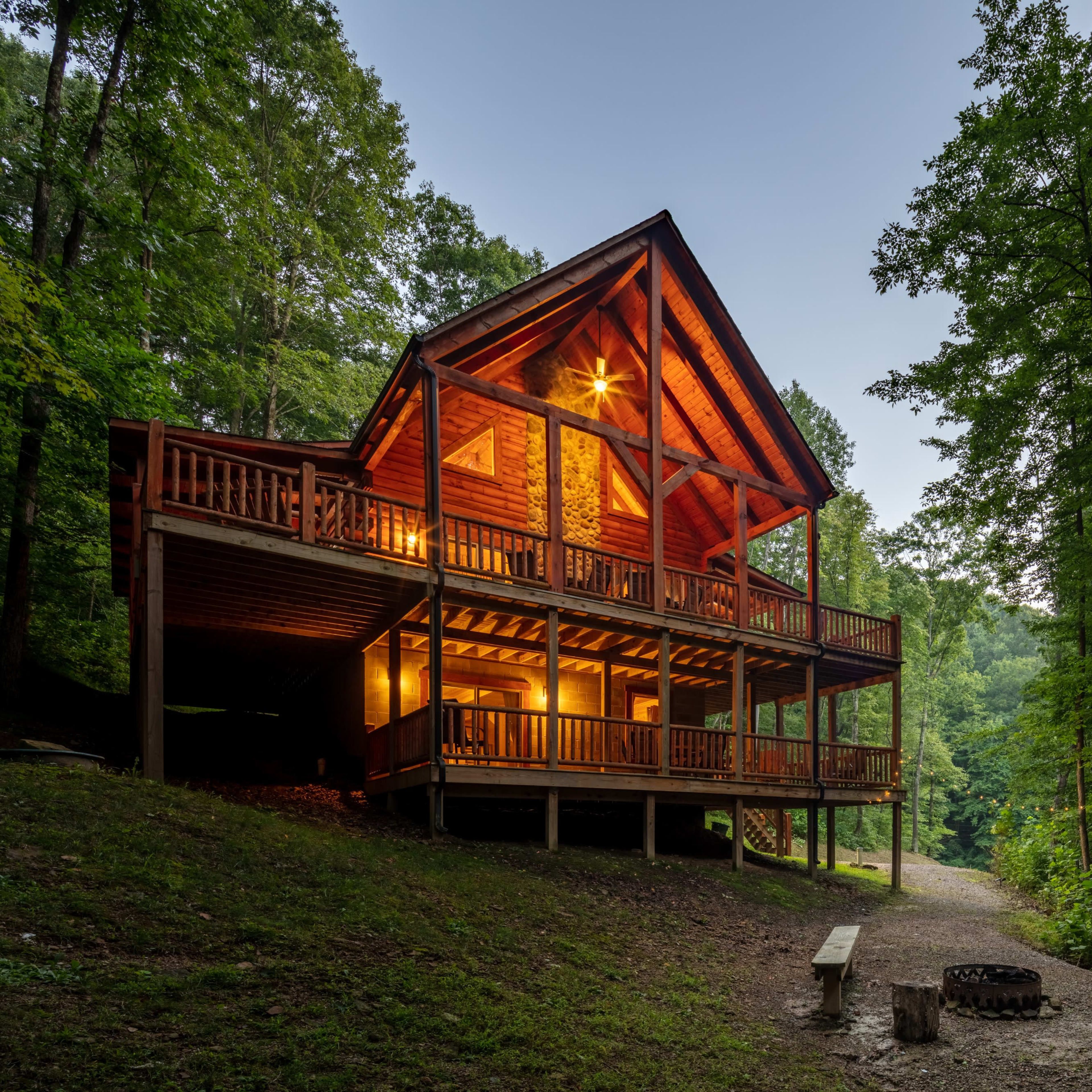 A large cabin with a wooden structure and balcony is surrounded by trees, with a pathway leading to a fire pit area.