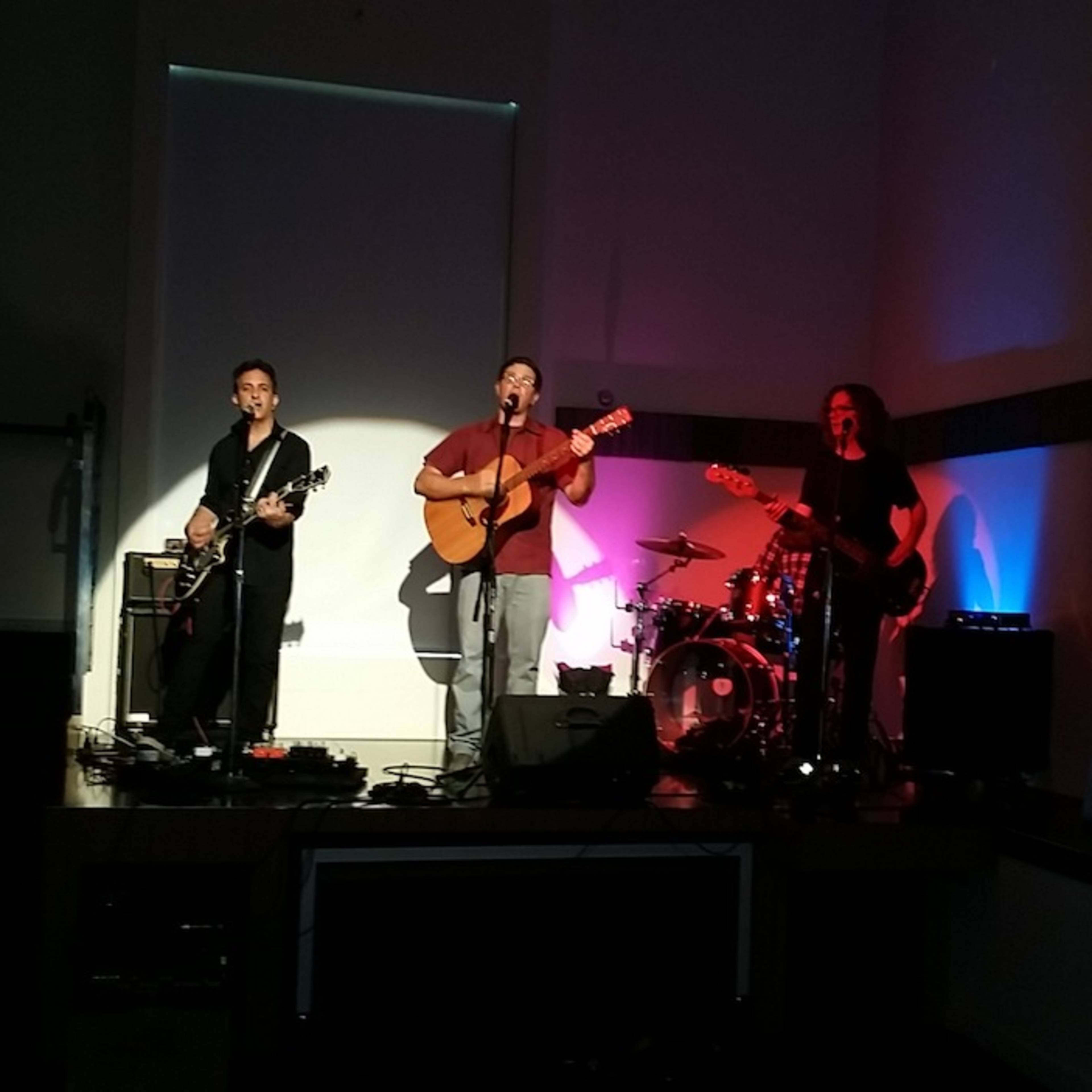 Three musicians perform on a small stage with instruments and colorful lighting.