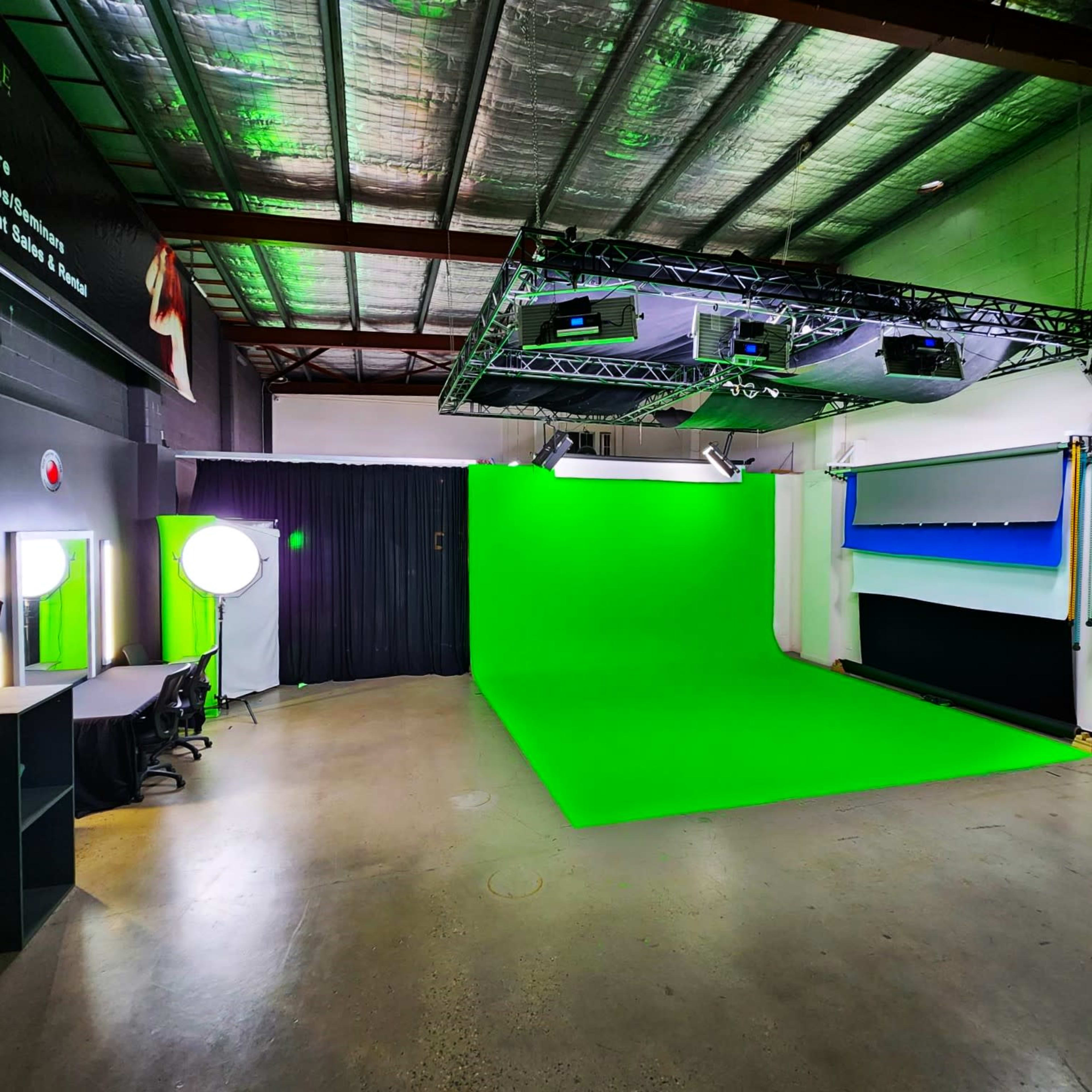 A green screen studio with bright green backdrop, professional lighting equipment, and a workspace area with a desk and chair.