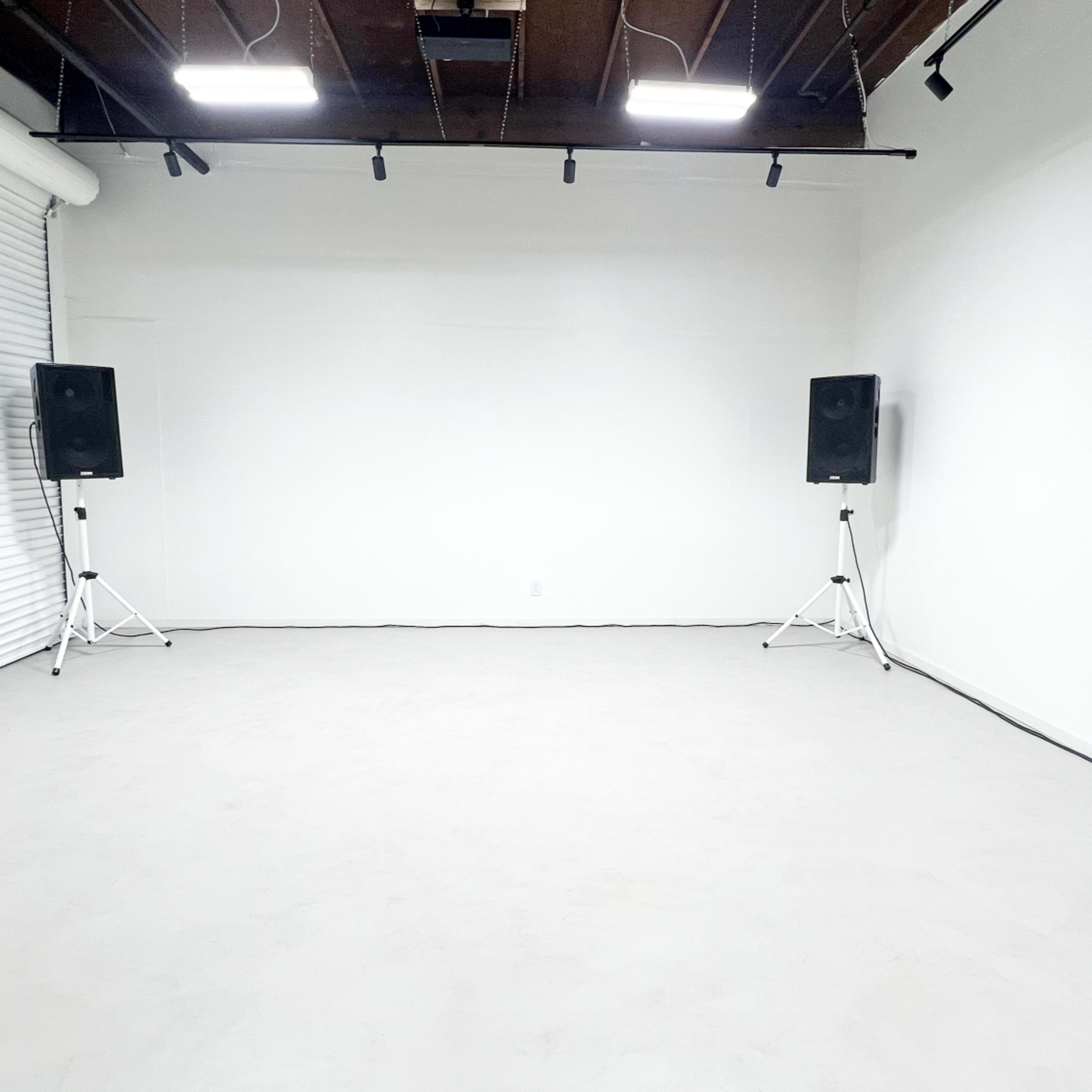 An empty room with white walls and a concrete floor, featuring two speakers mounted on stands against one wall.