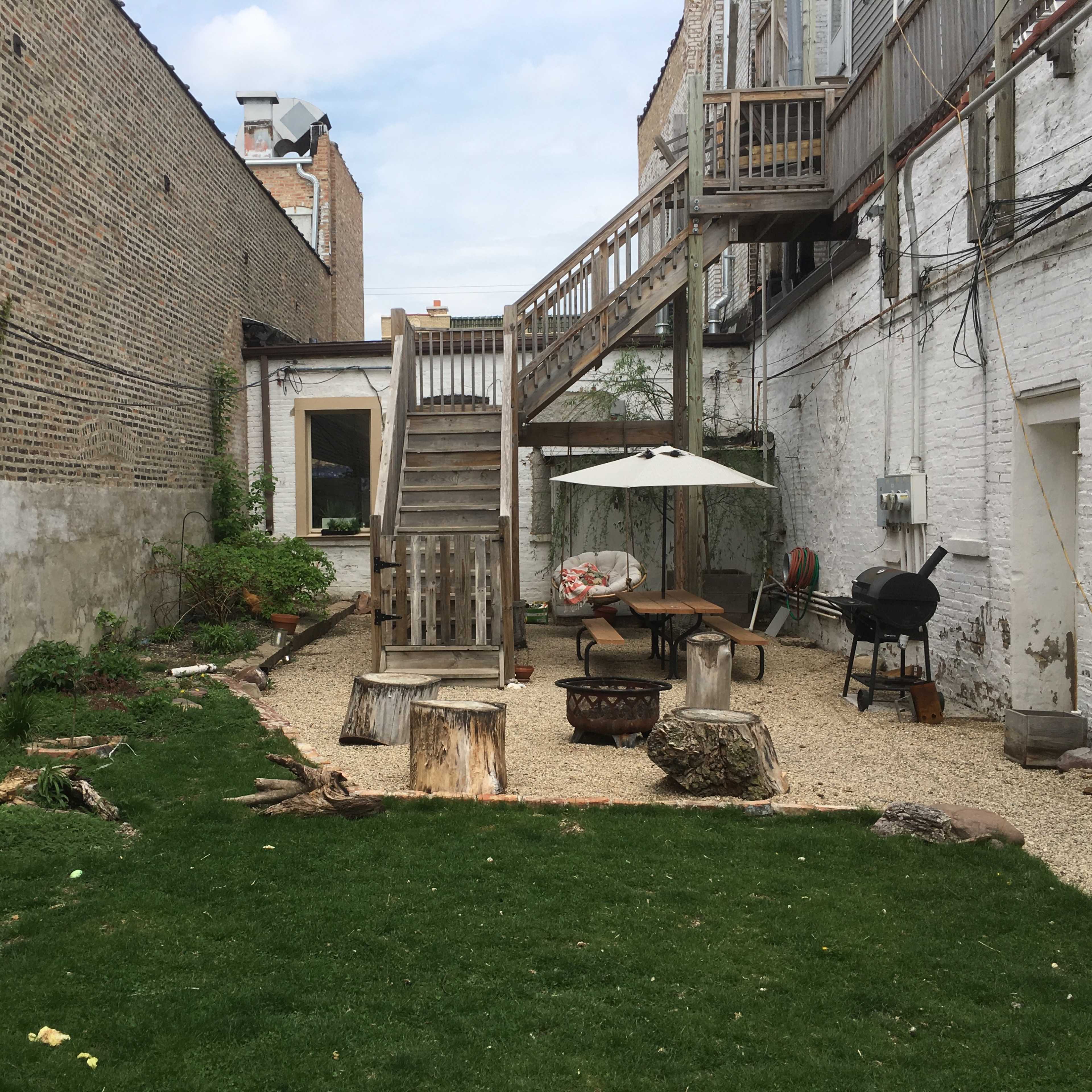 A gravel and grass backyard with a wooden deck, outdoor furniture, a fire pit, and a grill.