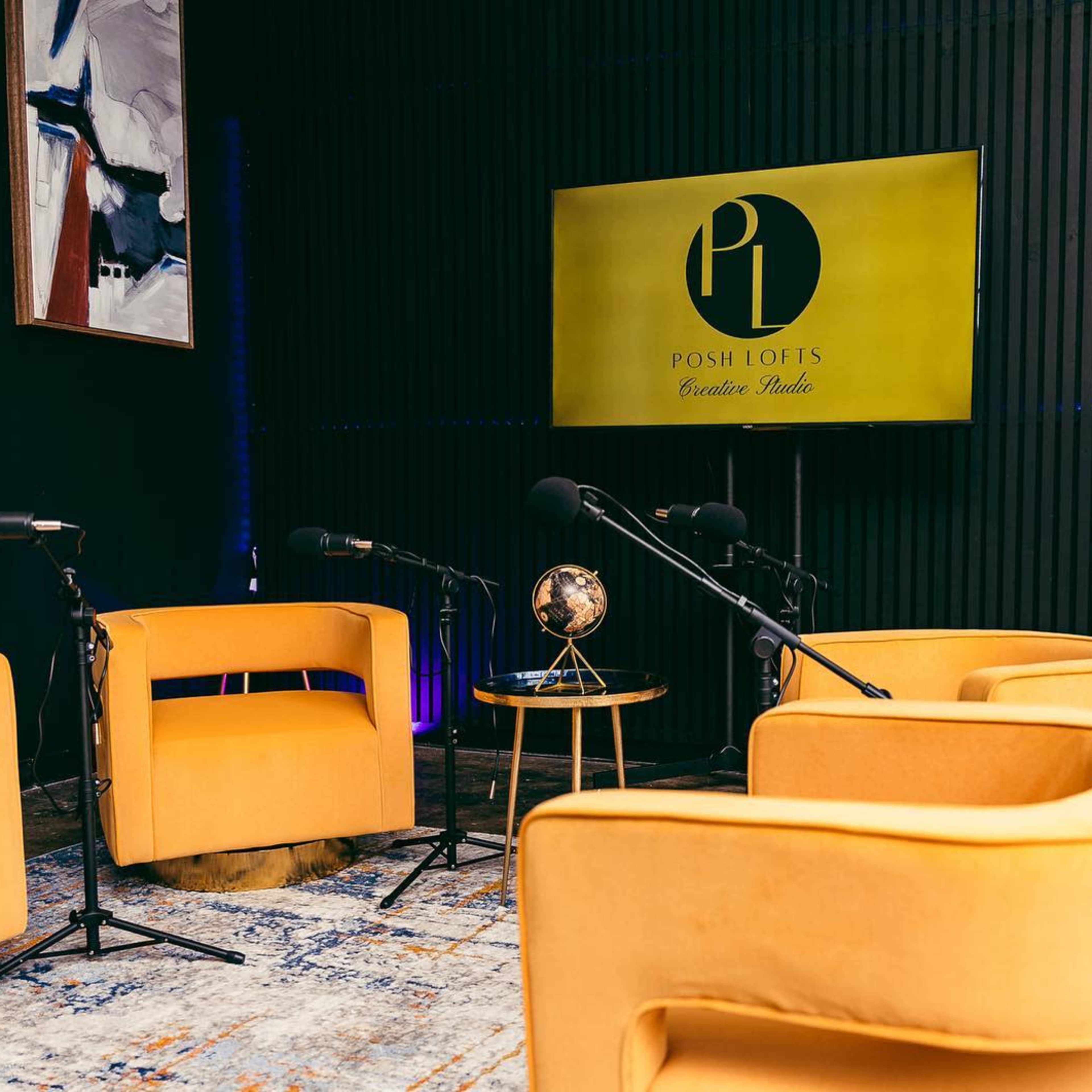A modern podcast setting with four orange chairs, a globe on a small table, and microphones arranged around a brightly lit screen displaying the logo "POSH LOFTS."