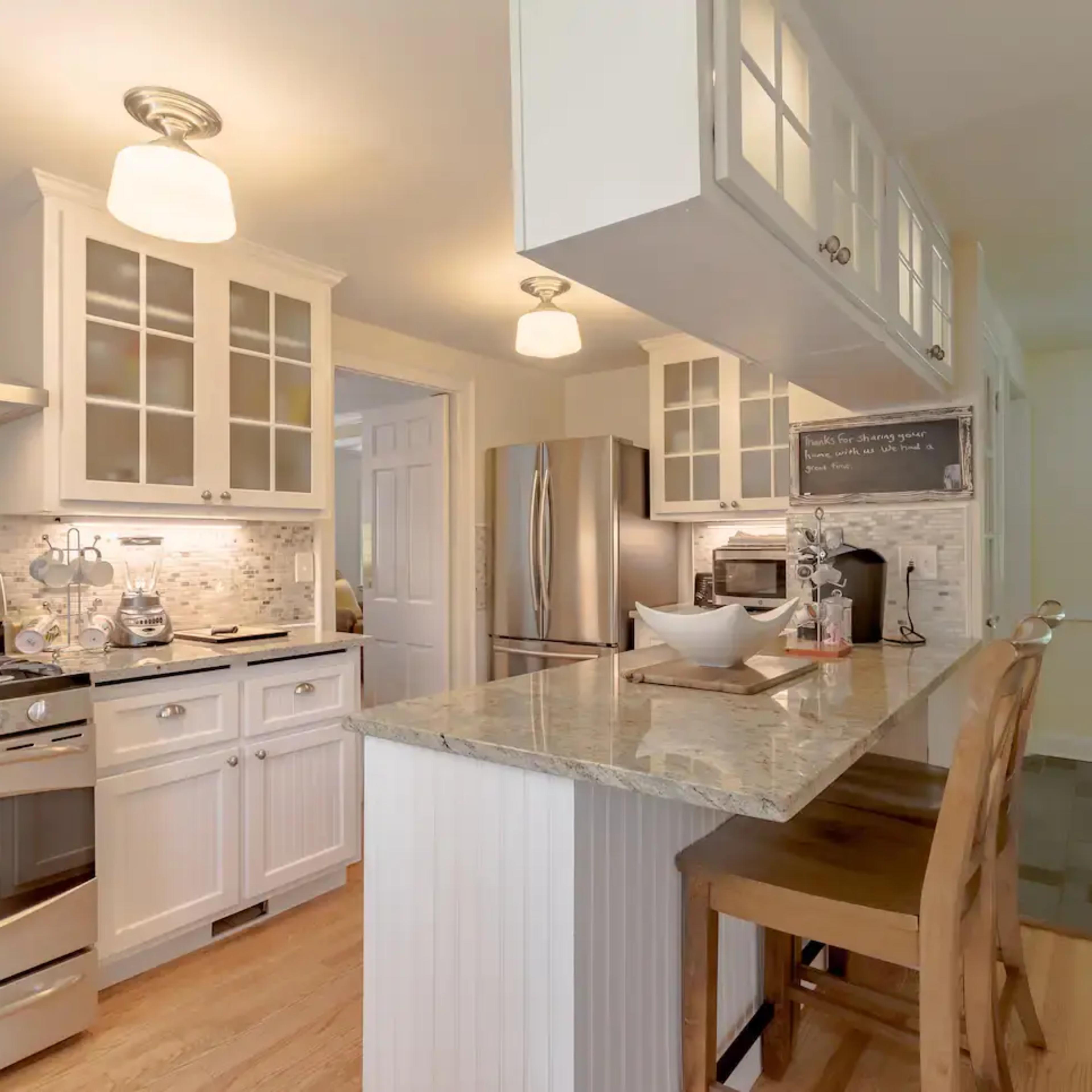 A modern kitchen with white cabinetry, a granite countertop, stainless steel appliances, and a dining area with a wooden chair.