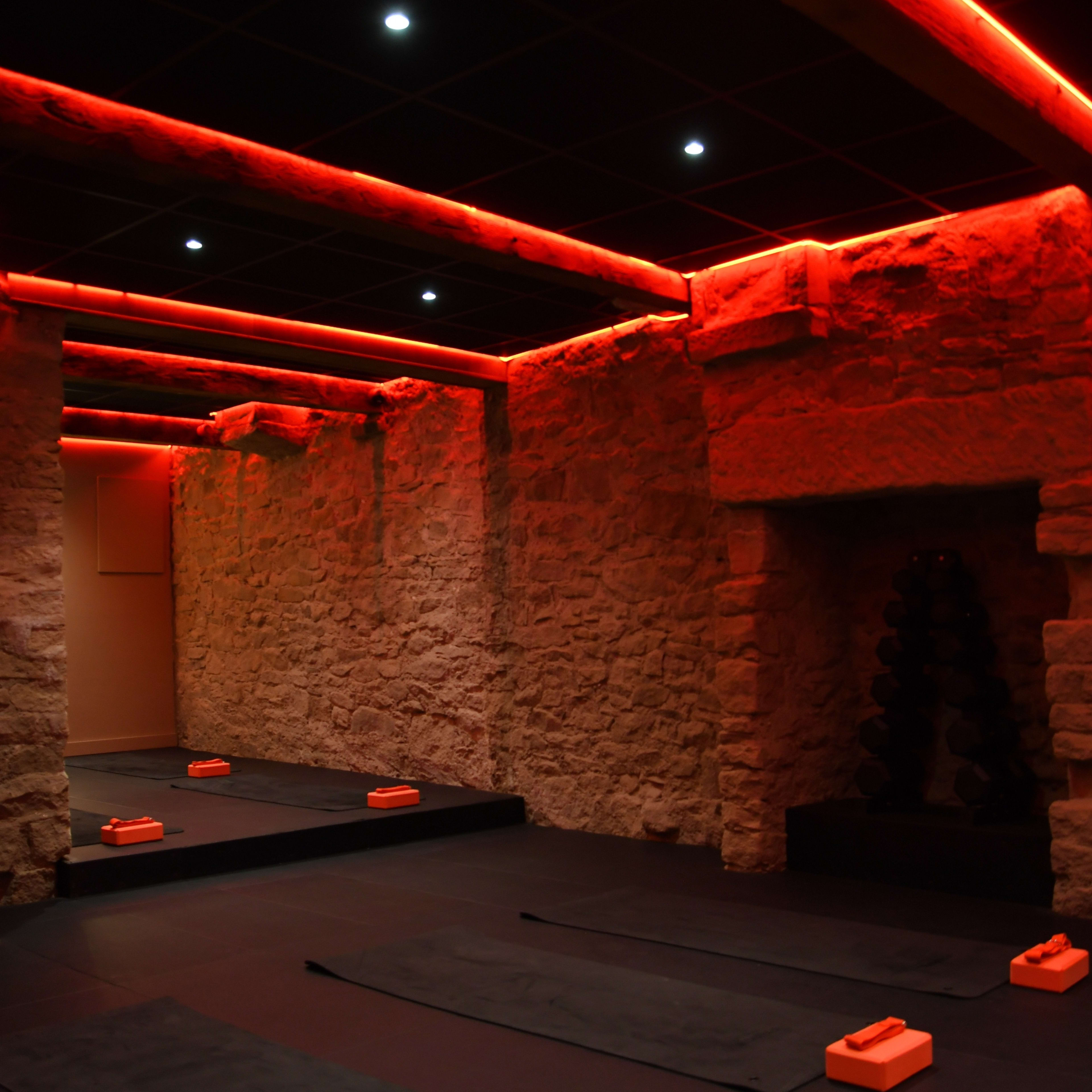 A dimly lit fitness studio featuring black yoga mats arranged on the floor, orange blocks positioned on each mat, and textured stone walls illuminated by red lighting.