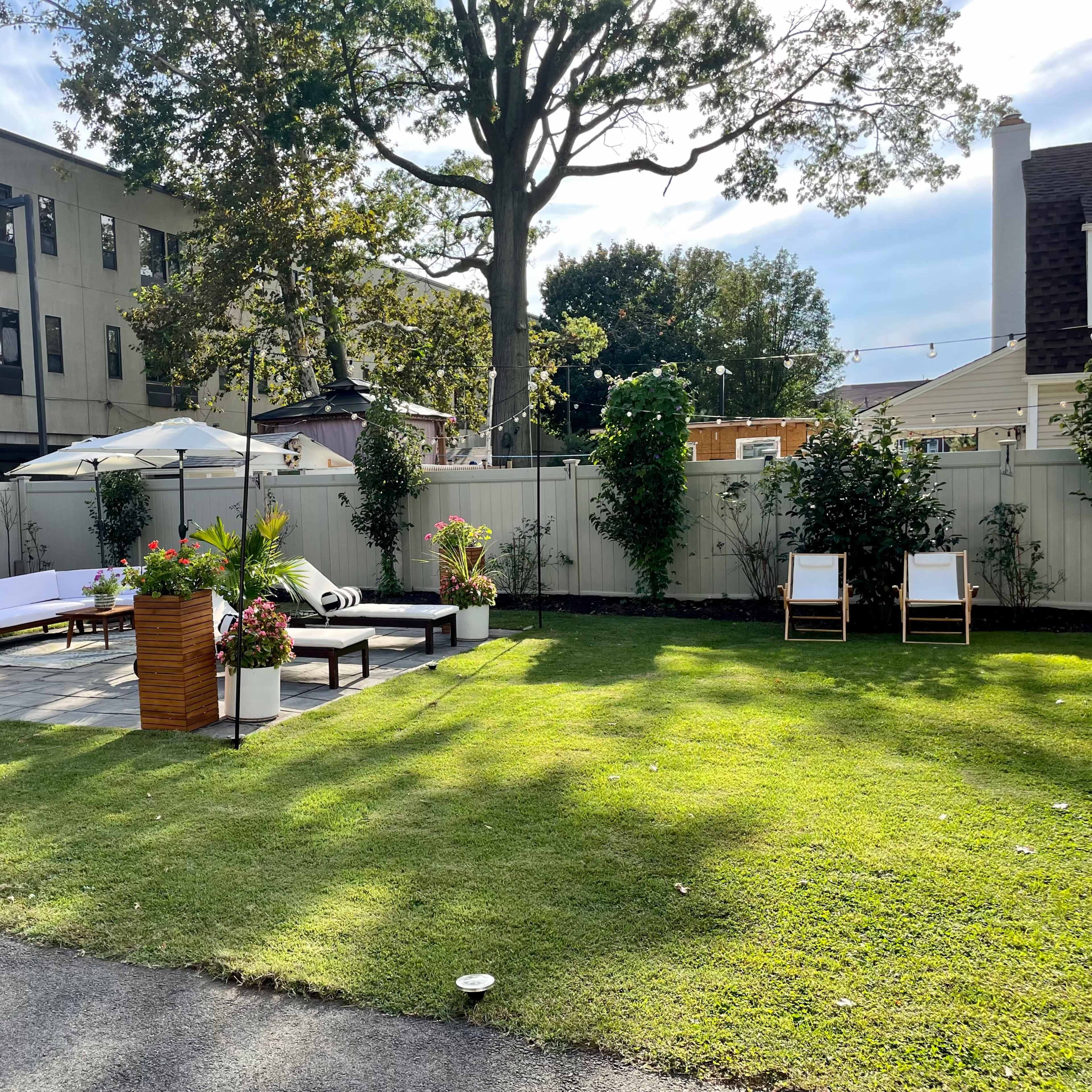 A landscaped outdoor space features lounge chairs, a shaded seating area, and decorative plants surrounded by a wooden fence.