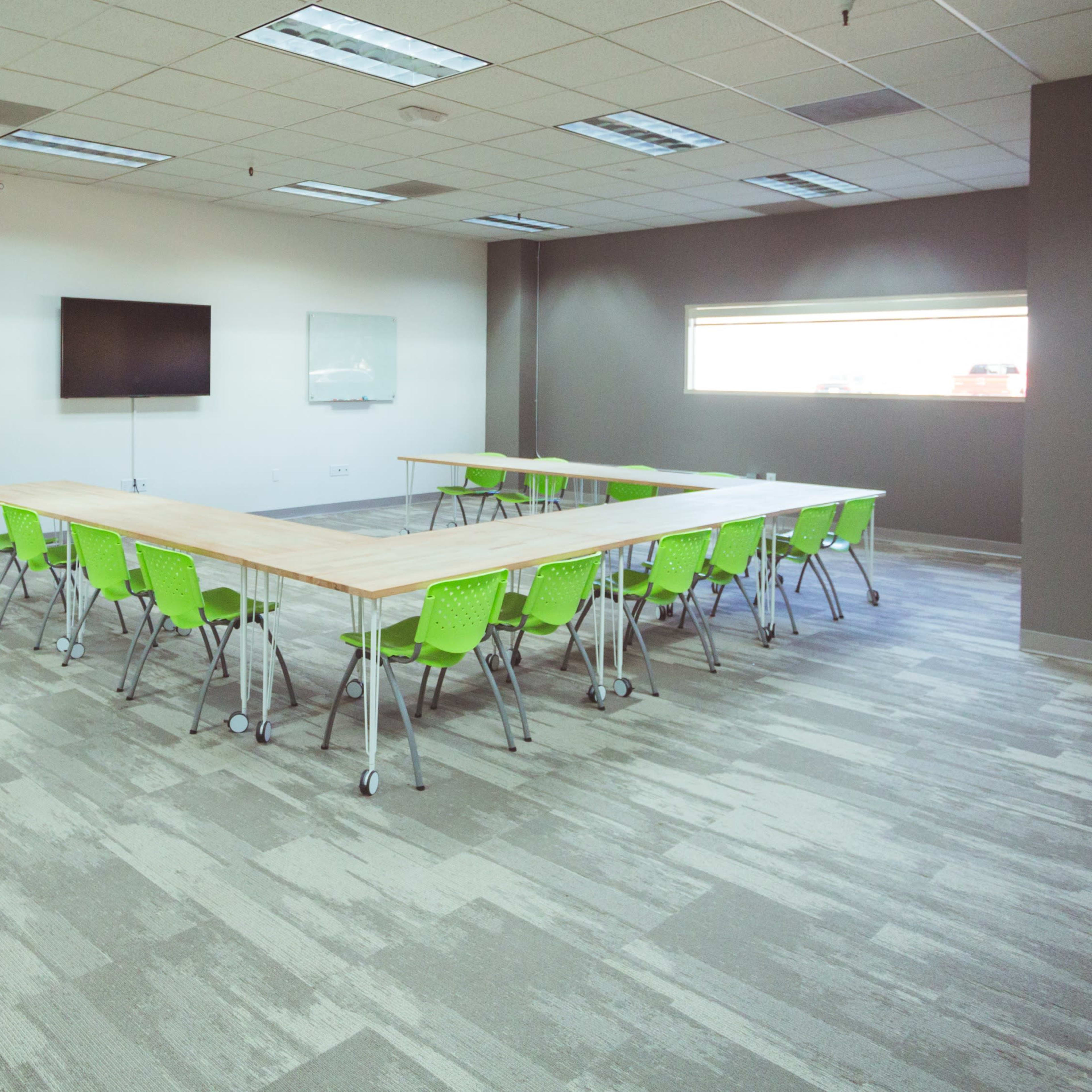 A modern conference room features a large wooden table surrounded by green chairs, with a wall displaying colorful artwork and a screen for presentations.