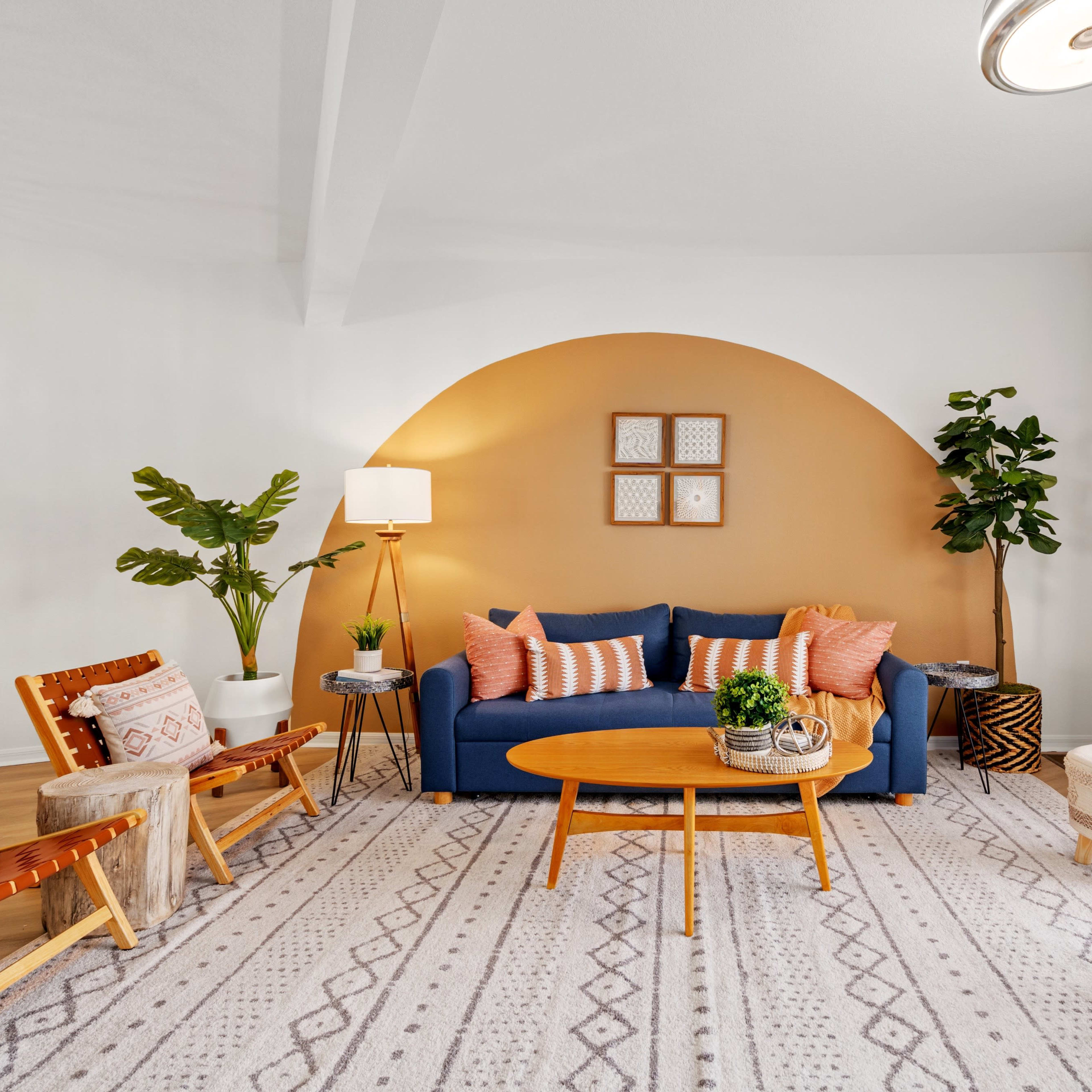 The living room features a blue sofa with orange and cream pillows, a round coffee table, and two wooden chairs on a patterned rug, set against a curved wall painted in a warm tone.