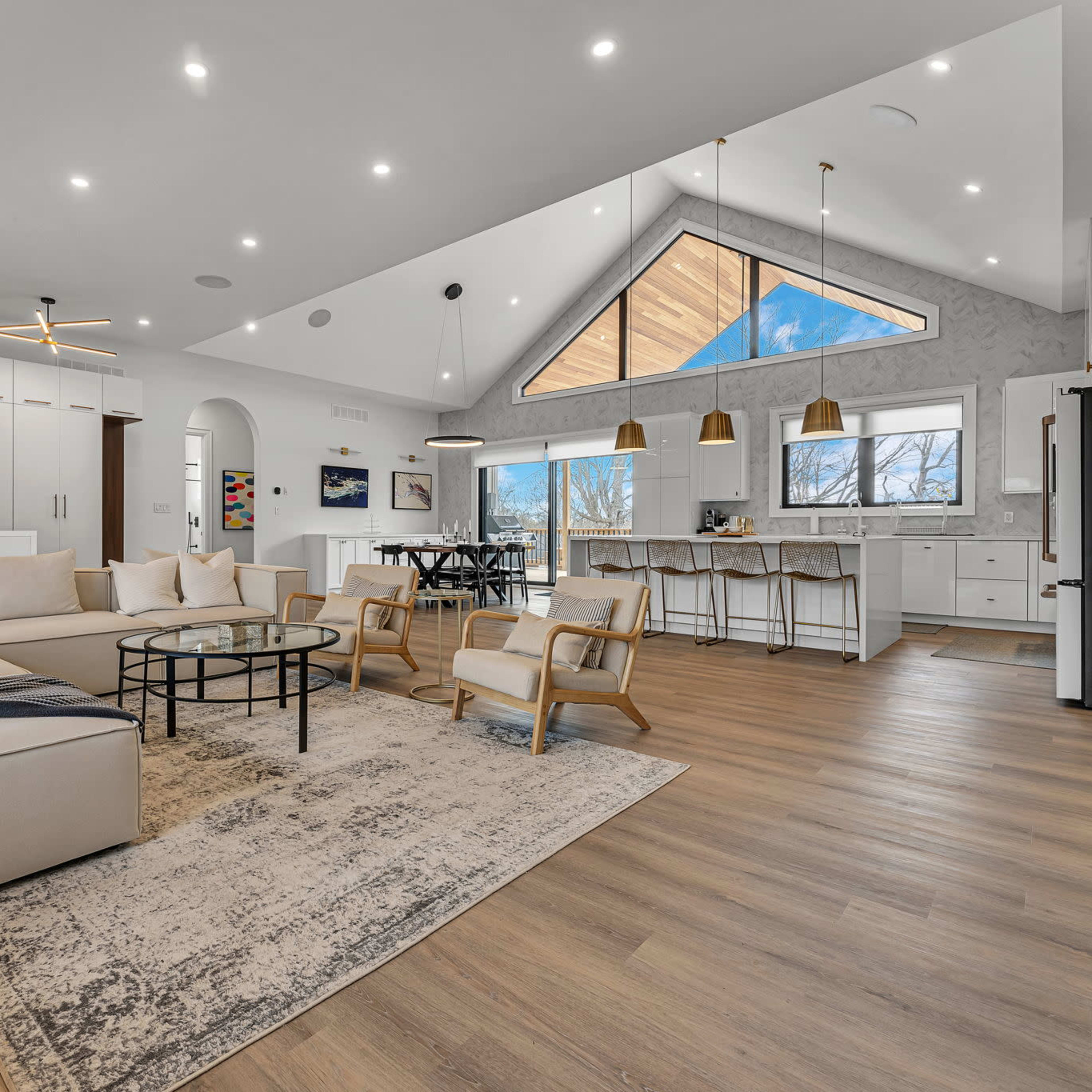 The modern living area features a sectional sofa and a circular coffee table, with an open layout leading to a dining space and kitchen, highlighted by large windows and pendant lighting.