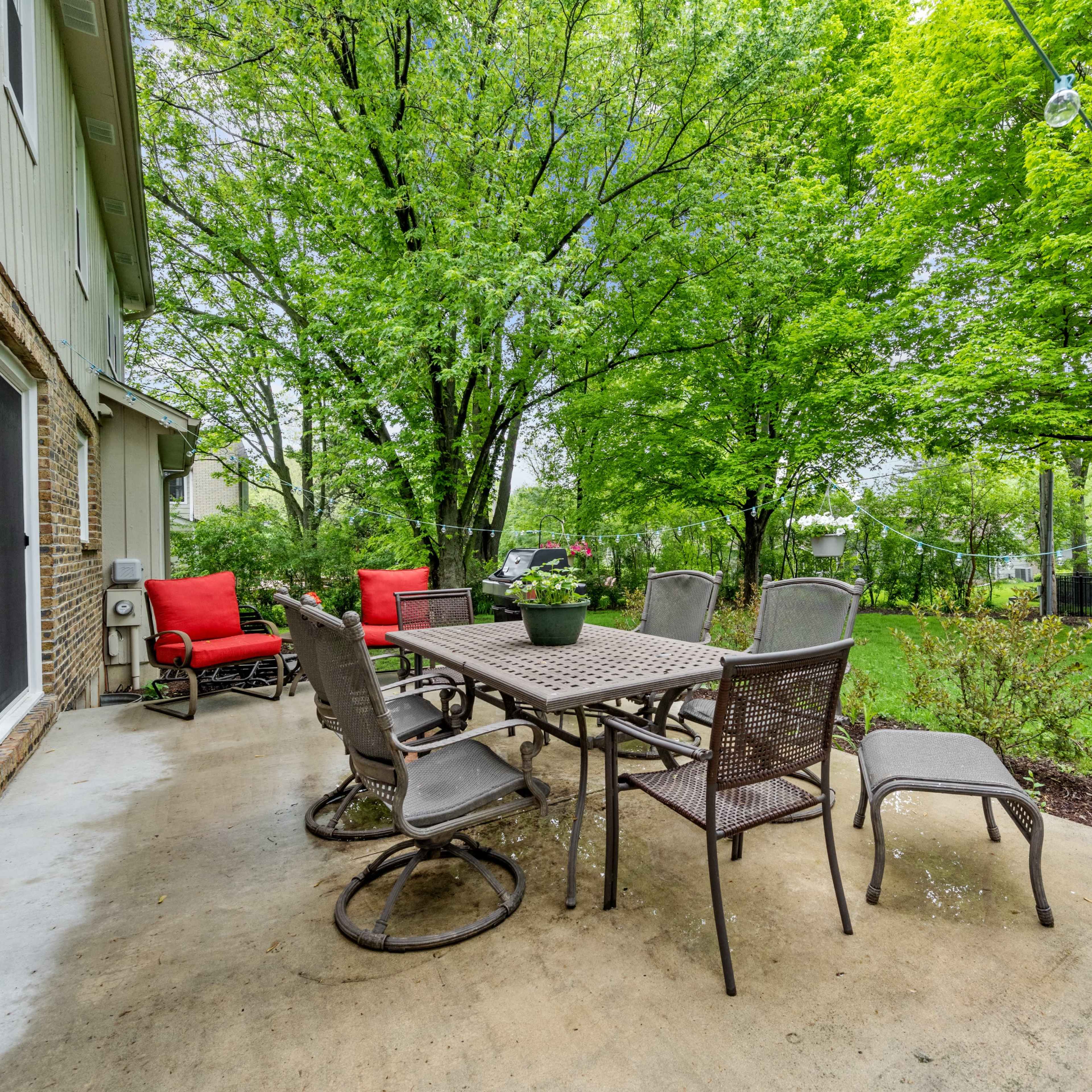 A patio features a rectangular table surrounded by chairs, with red armchairs positioned nearby, all set against a backdrop of lush green trees.