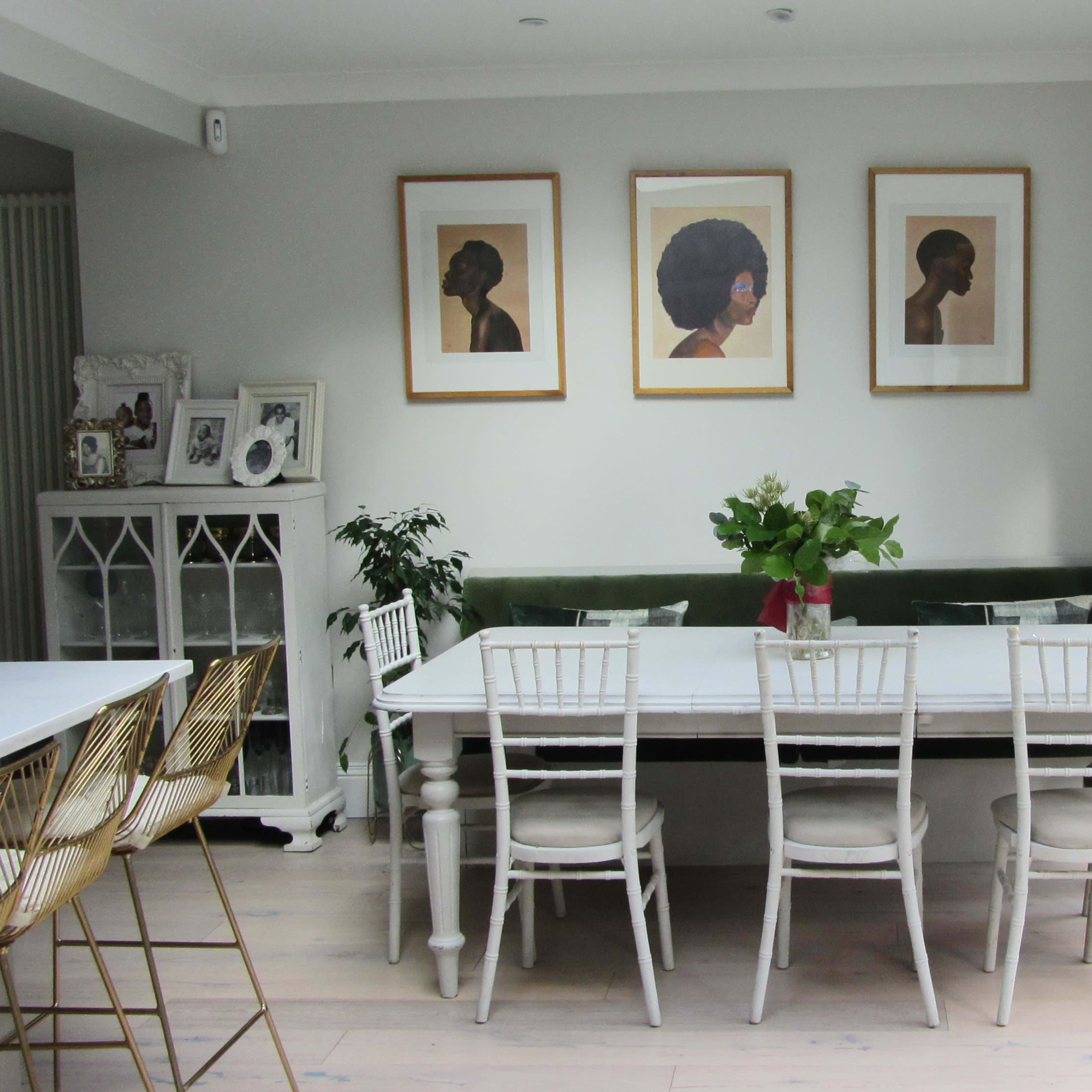 A dining area features a long white table surrounded by several white chairs, with framed portraits on the wall and a green banquette along one side.