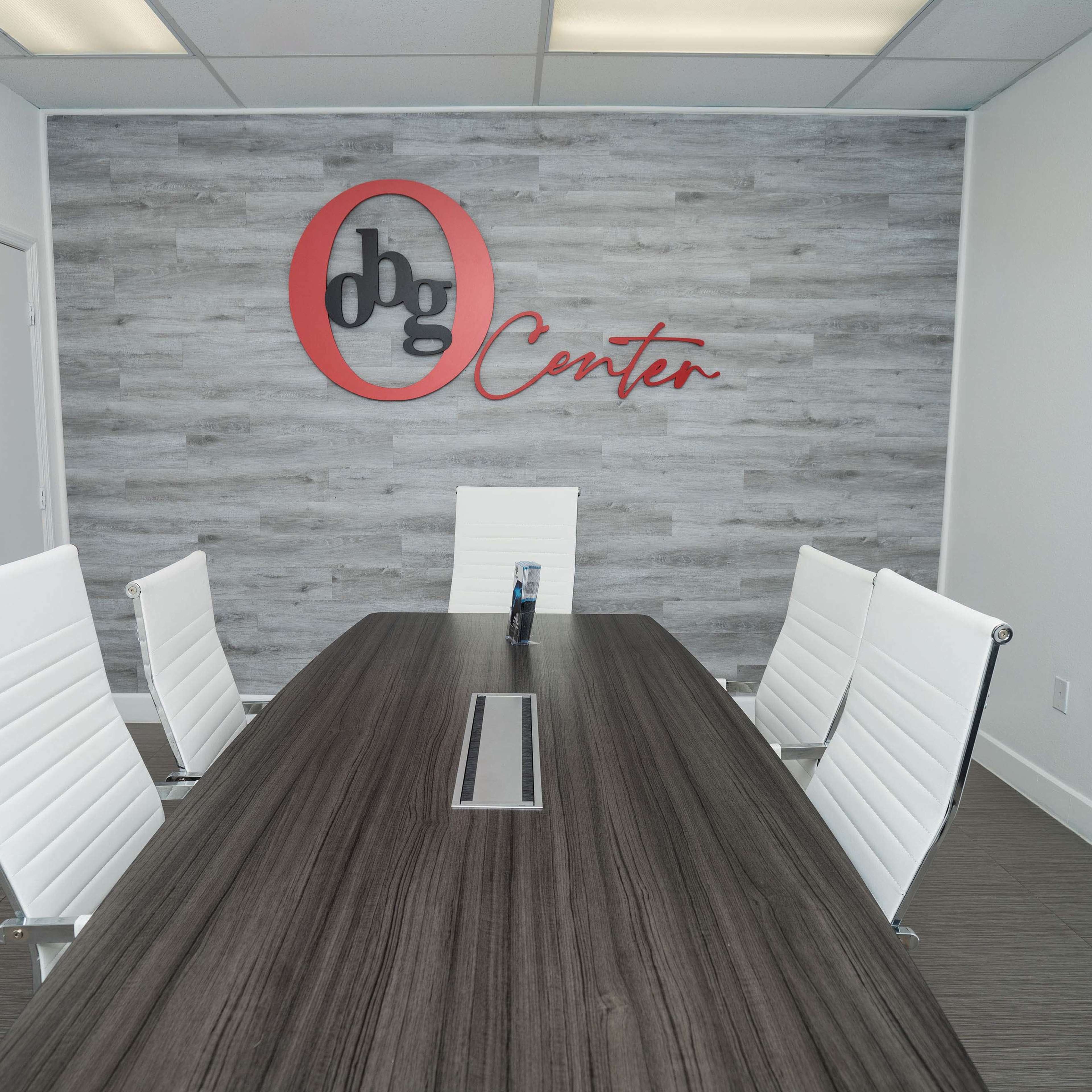 A conference room features a wooden table with a central cable management slot, surrounded by four white chairs and a branded wall with the text "dbg Center."