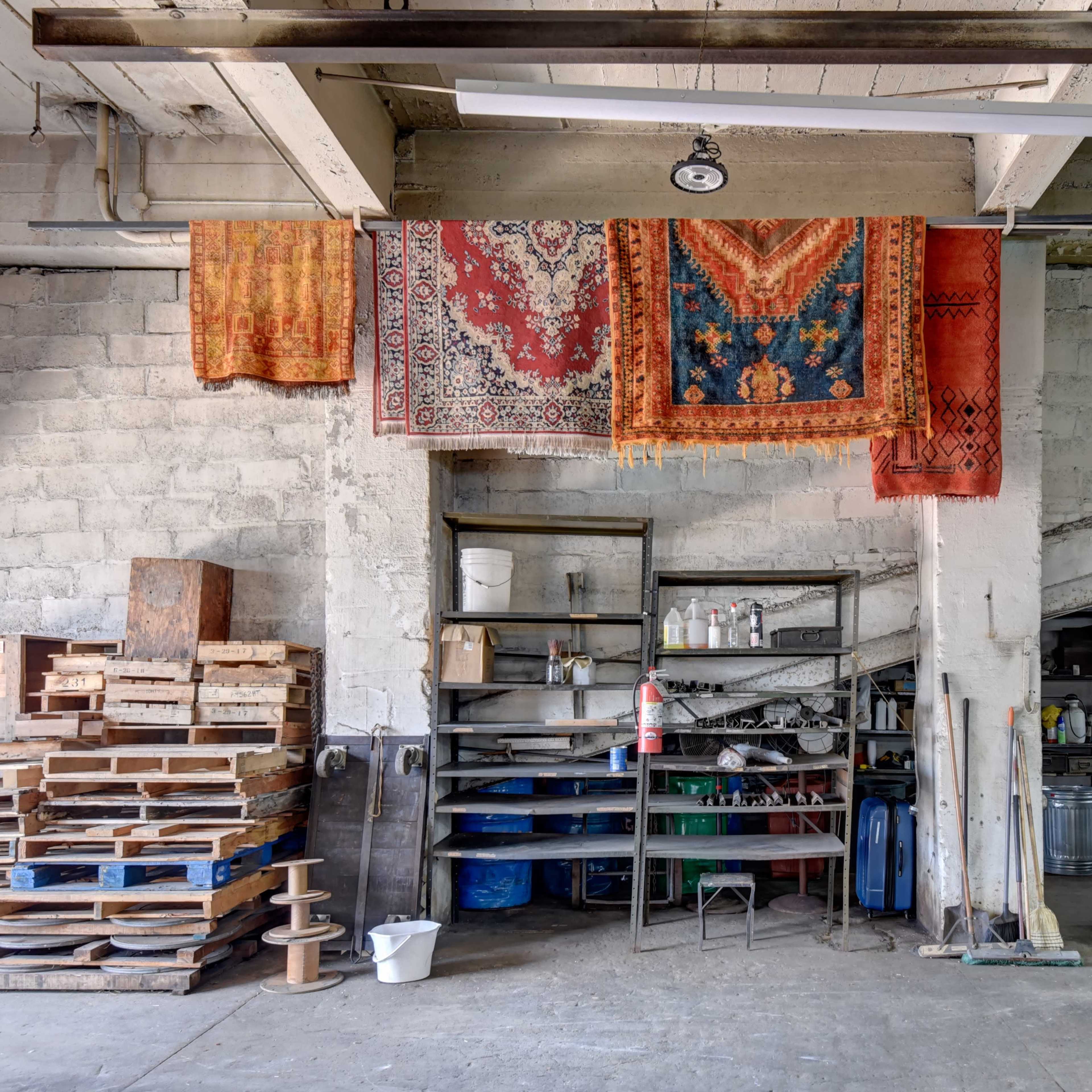 A cluttered workshop features stacked wooden pallets, shelves filled with various tools and materials, and colorful rugs hanging on the walls.