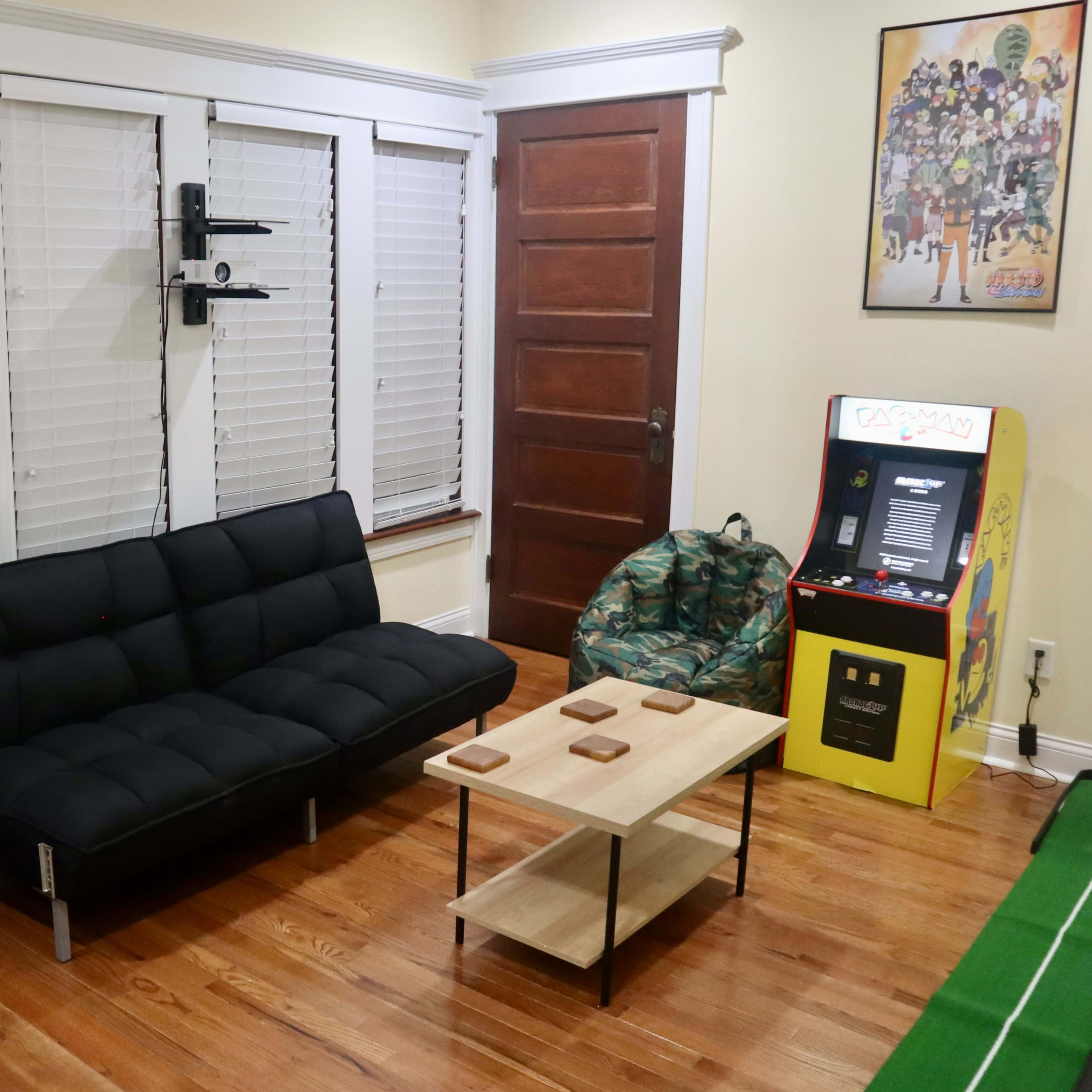 A living room featuring a black sofa, two camouflaged bean bags, a wooden coffee table, and a vintage arcade game machine in a brightly colored cabinet.