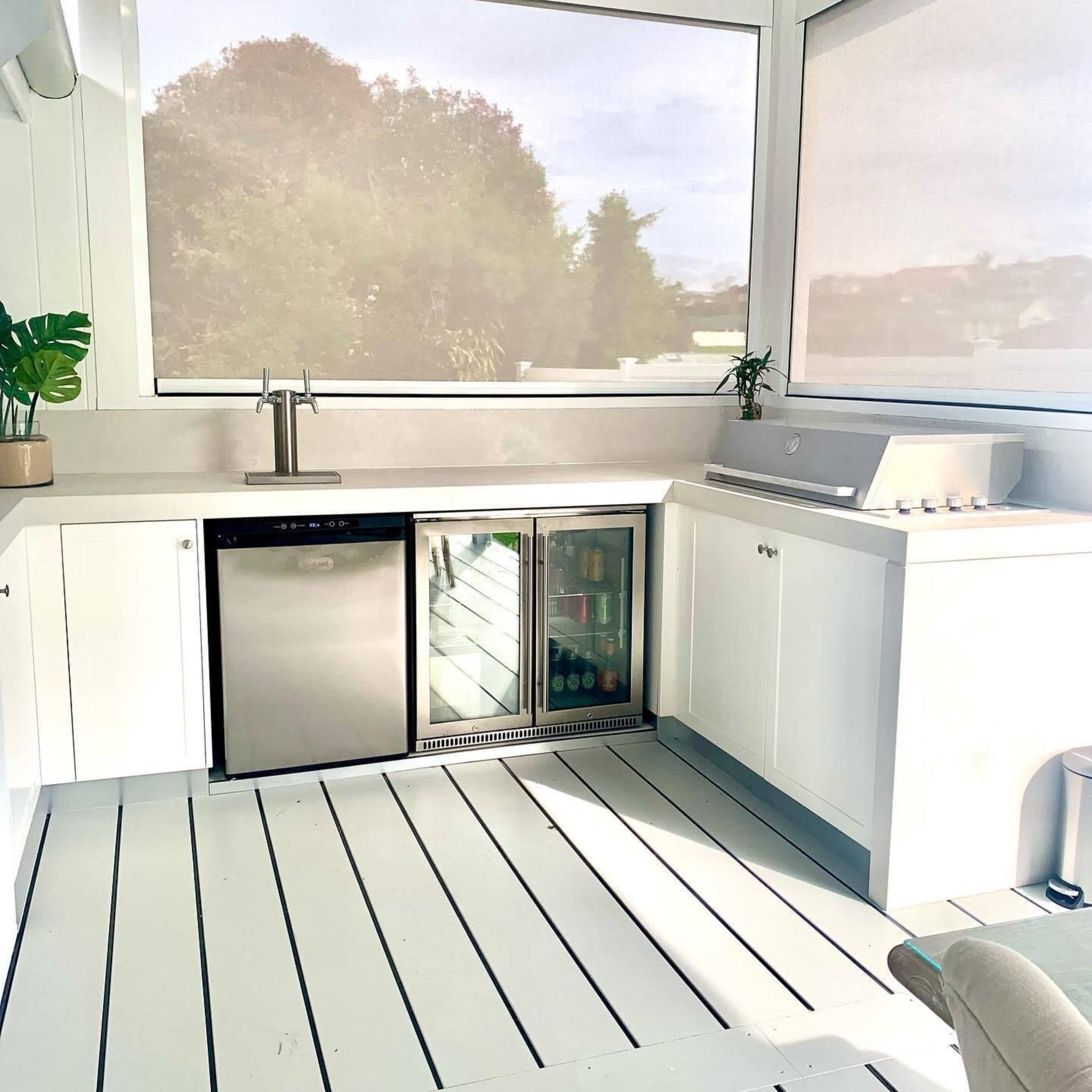 A modern outdoor kitchen with a sink, a refrigerator, and a grill, all within a light-colored, enclosed space featuring large windows.