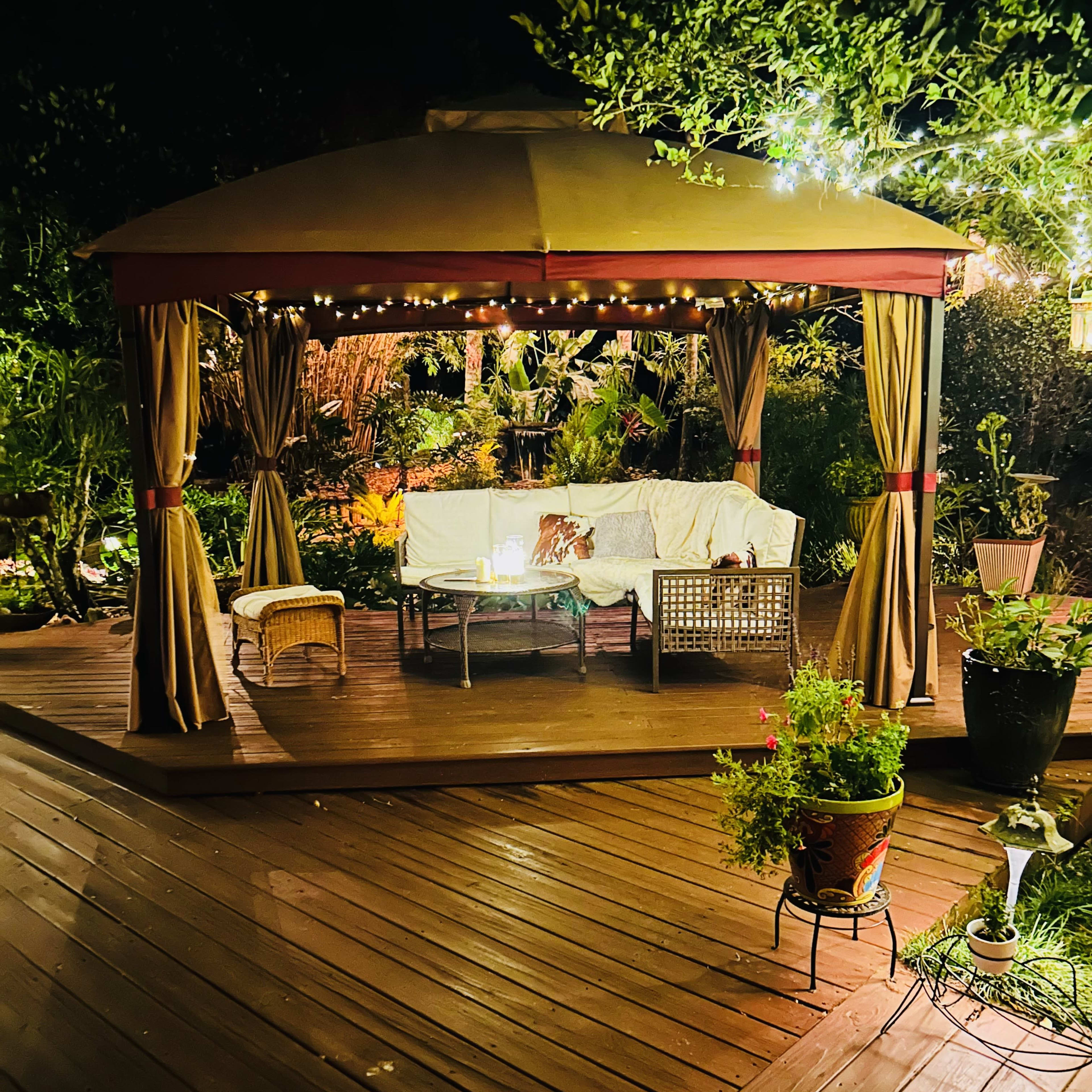 A gazebo surrounded by lush greenery is illuminated by string lights, featuring a seating area with a couch and potted plants on a wooden deck.