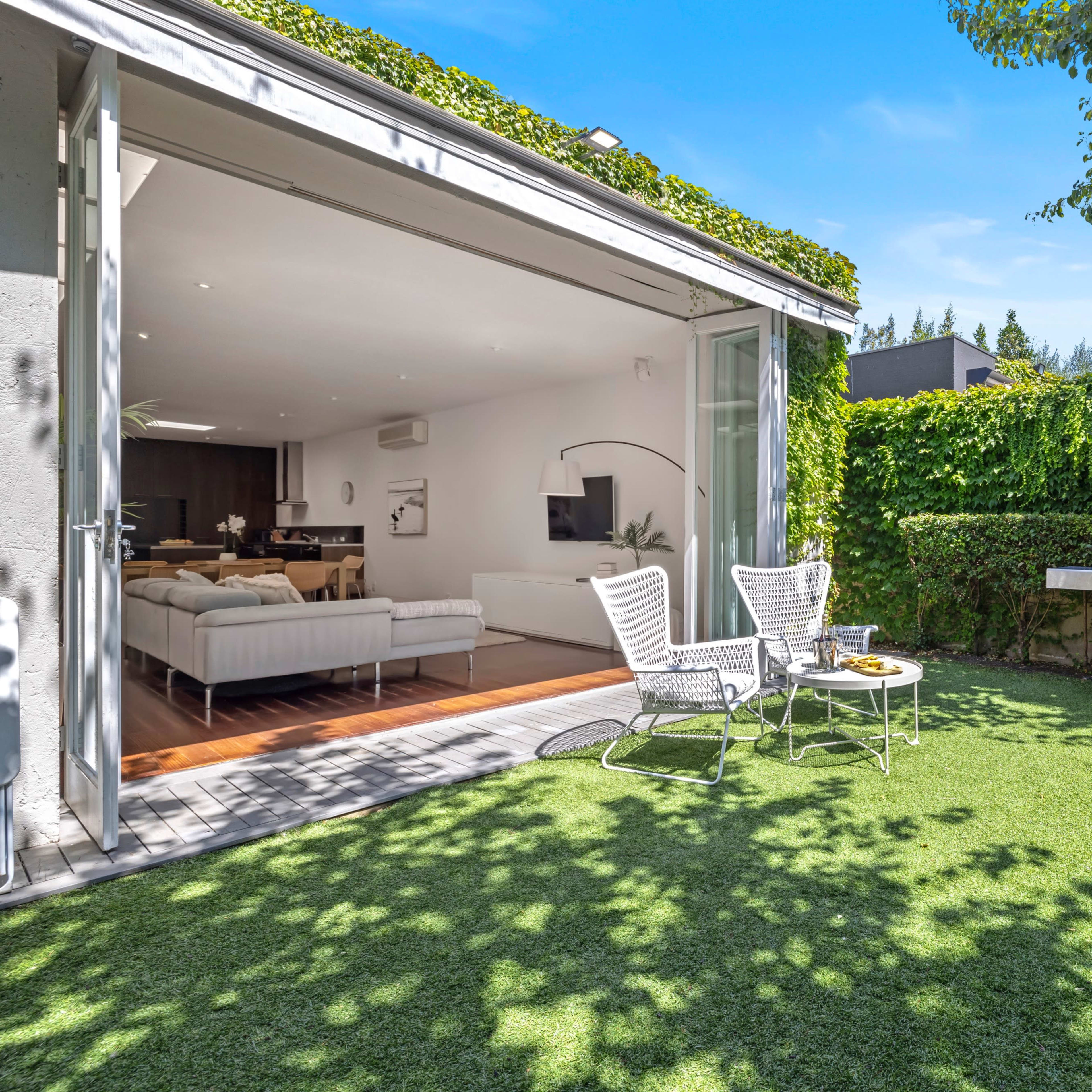 A modern backyard features two chairs, a small table, and a stainless steel grill surrounded by lush green grass and hedges.