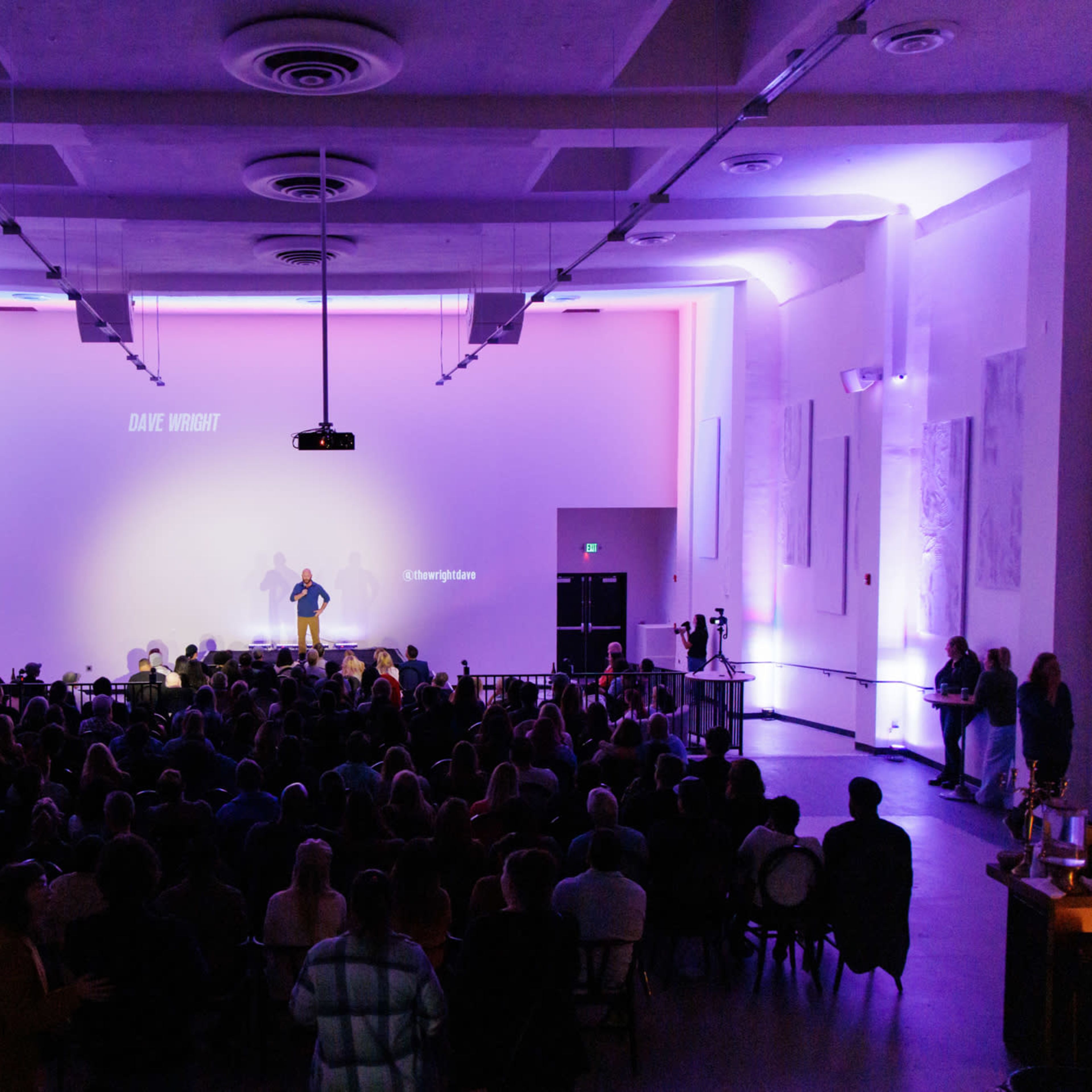 A speaker stands on stage in a well-lit auditorium filled with an audience, with a large screen displaying text in the background.