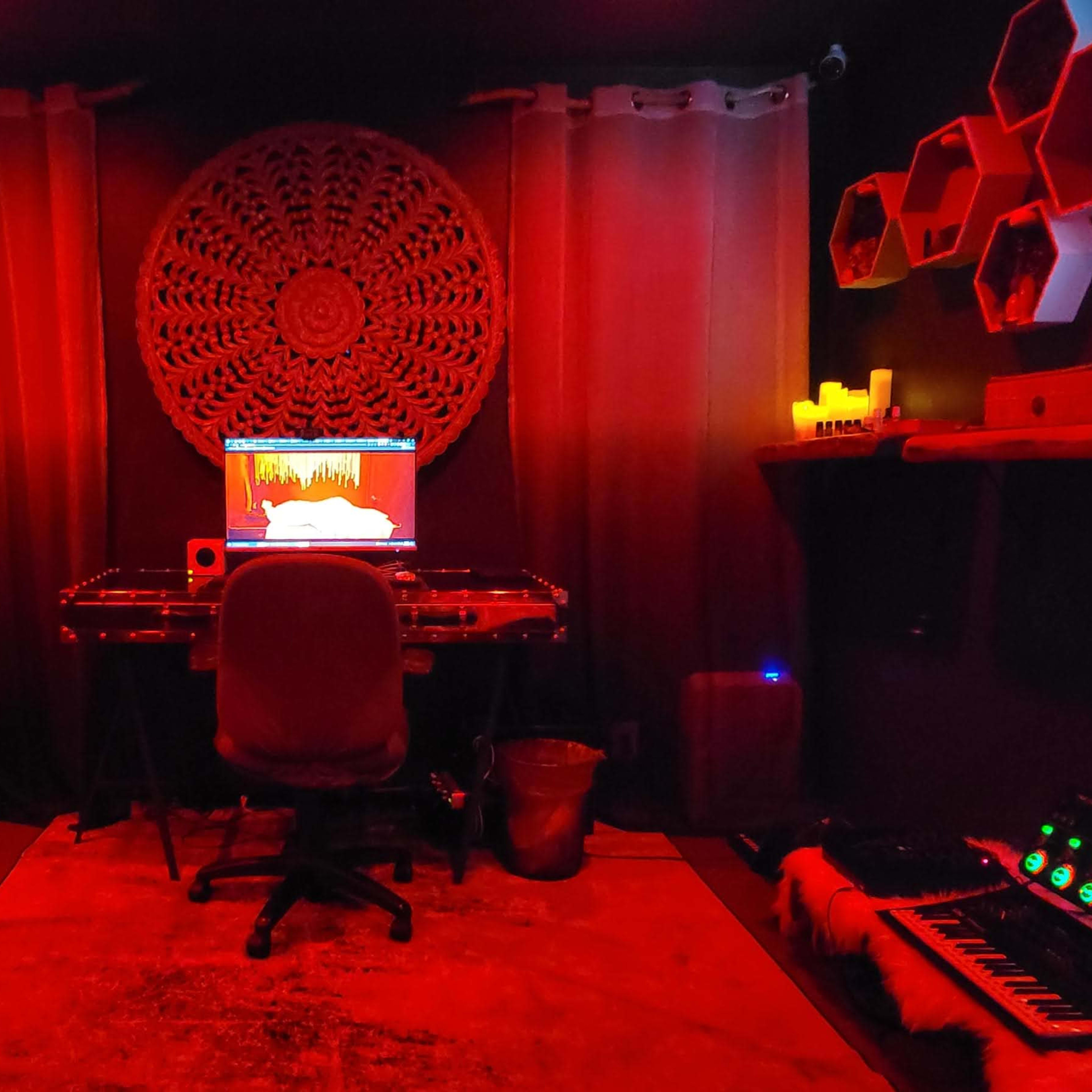 A music production studio features a computer setup with a glowing screen, surrounded by a red-lit room with decorative elements and a couch.
