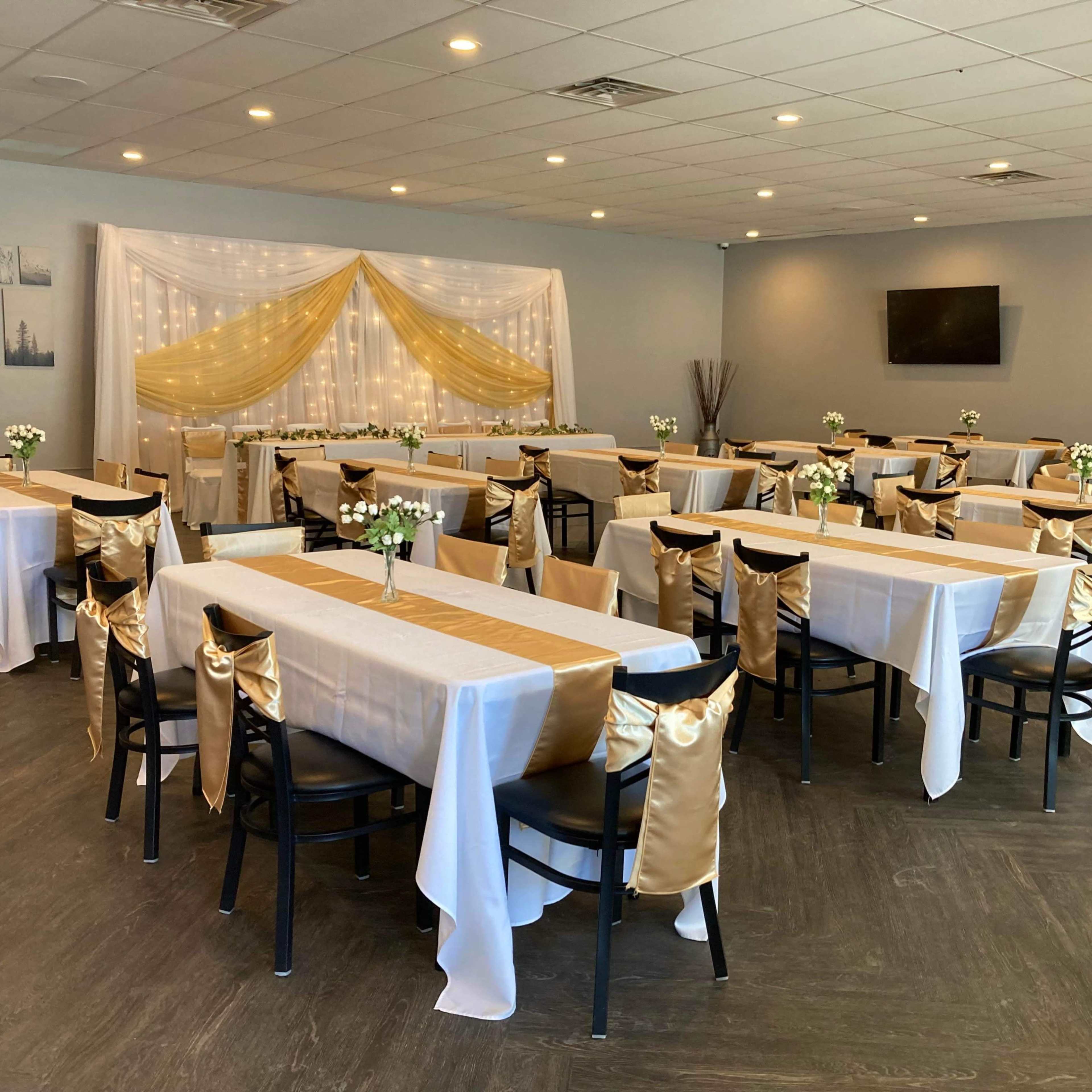 A banquet hall set up with several tables covered in white and gold tablecloths, arranged with small flower centerpieces.