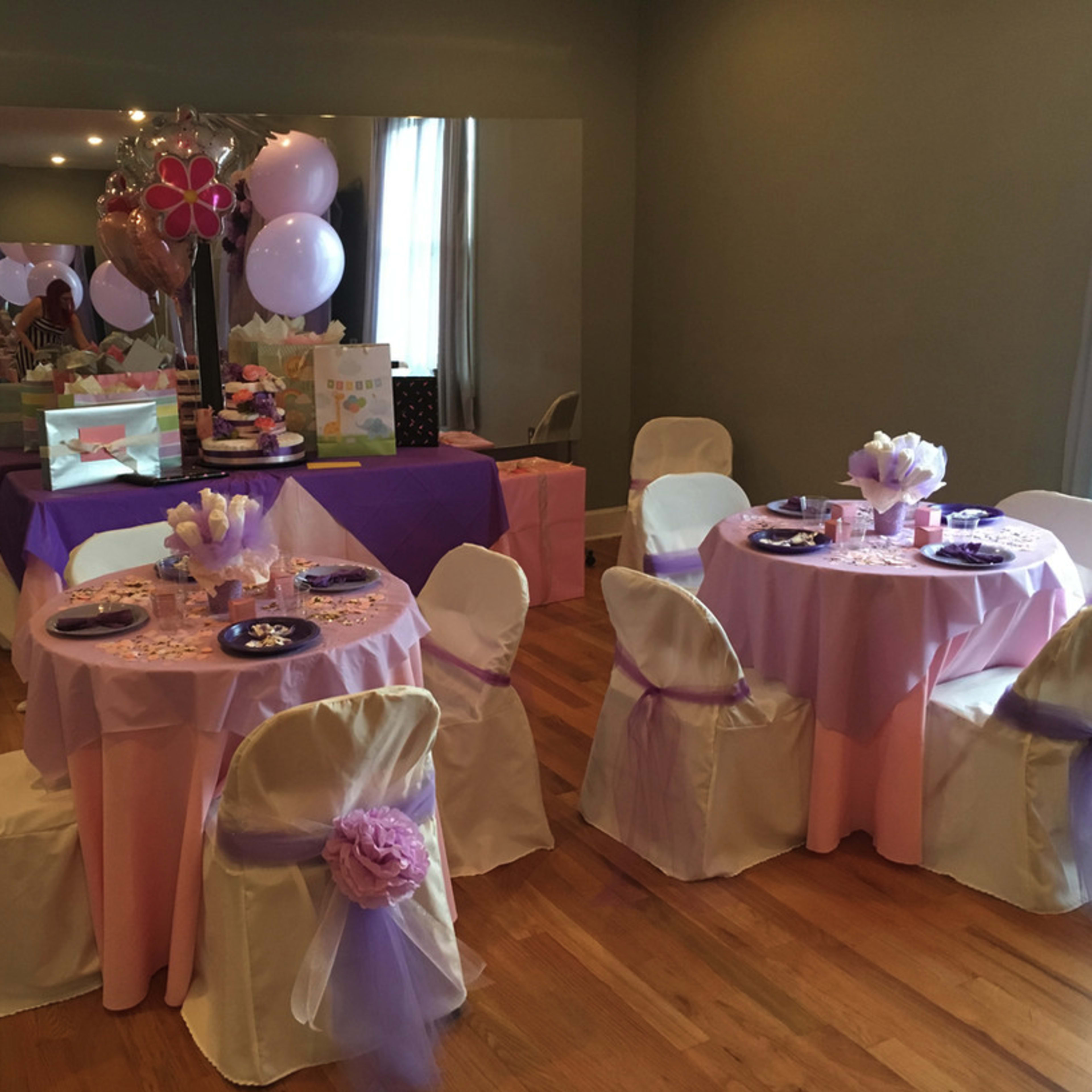 A decorated party space features tables set with pink and purple tableware, surrounded by chairs draped in white sashes, and accented by balloons and a cake display.