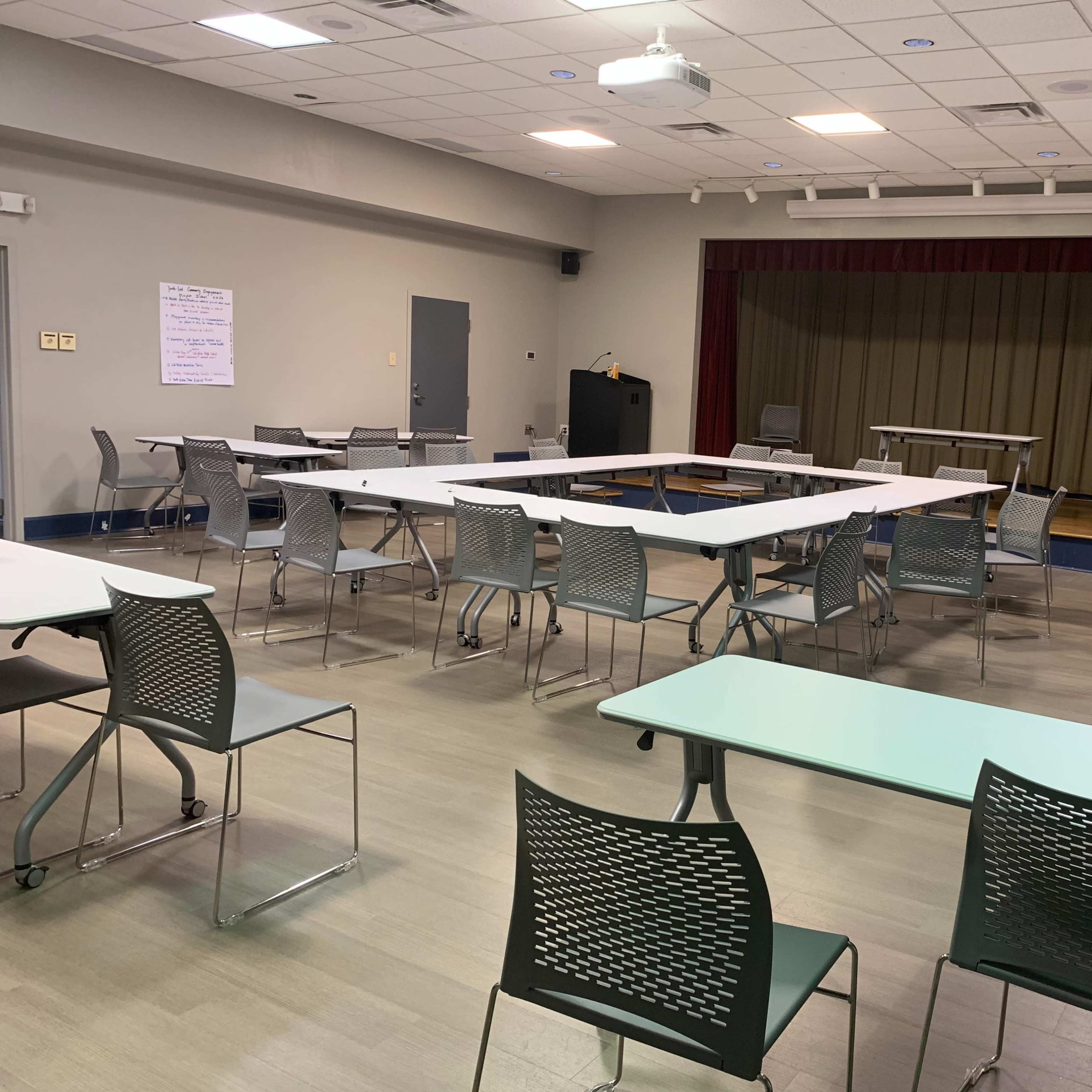 A well-lit meeting room with several tables and chairs arranged in a sparse layout, facing a small stage area.