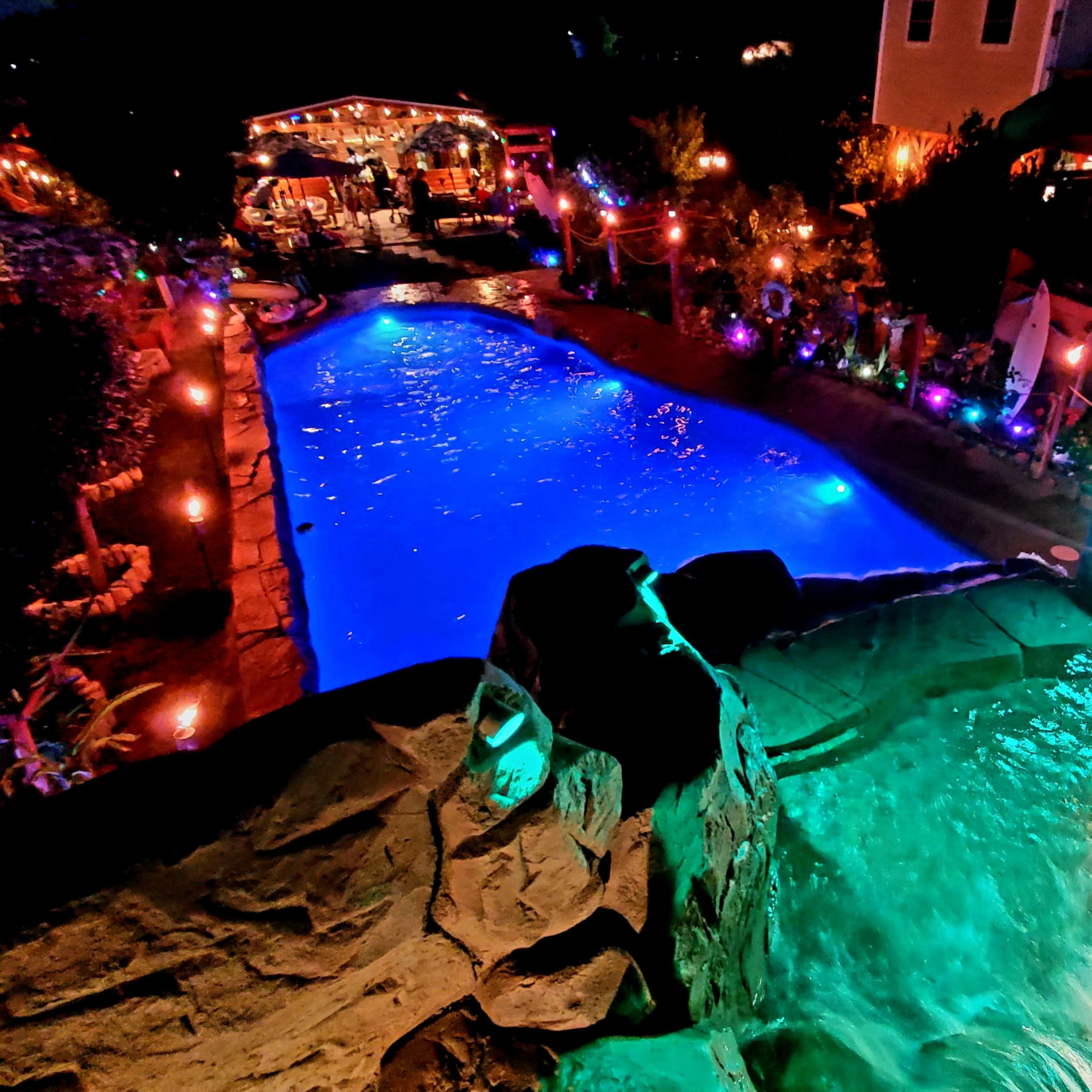 A lit outdoor pool area with colorful lights, a waterfall feature, and a gathering space in the background.