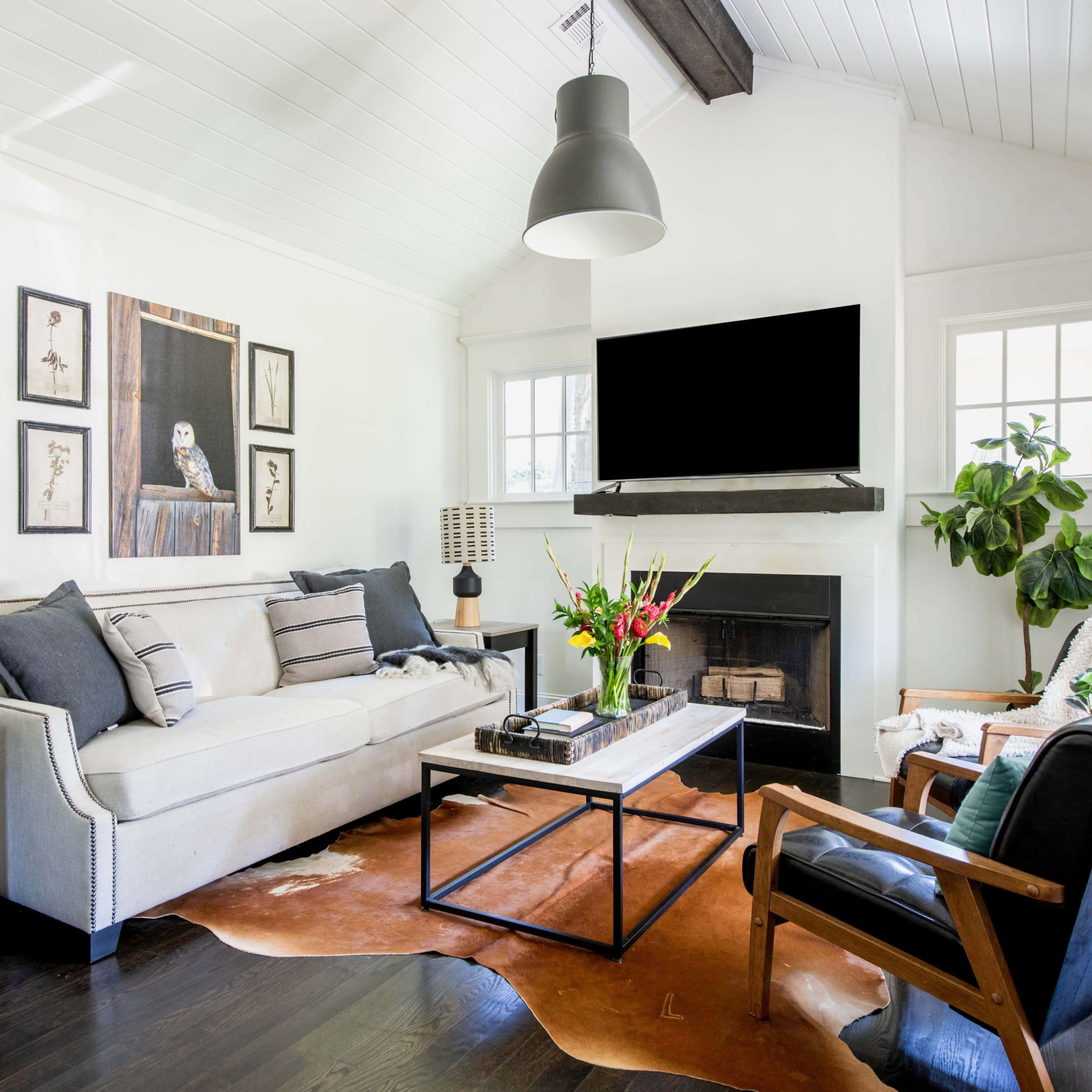 A modern living room featuring a beige sofa, a black coffee table, and a large television mounted above a fireplace, with accent chairs and decorative plants.