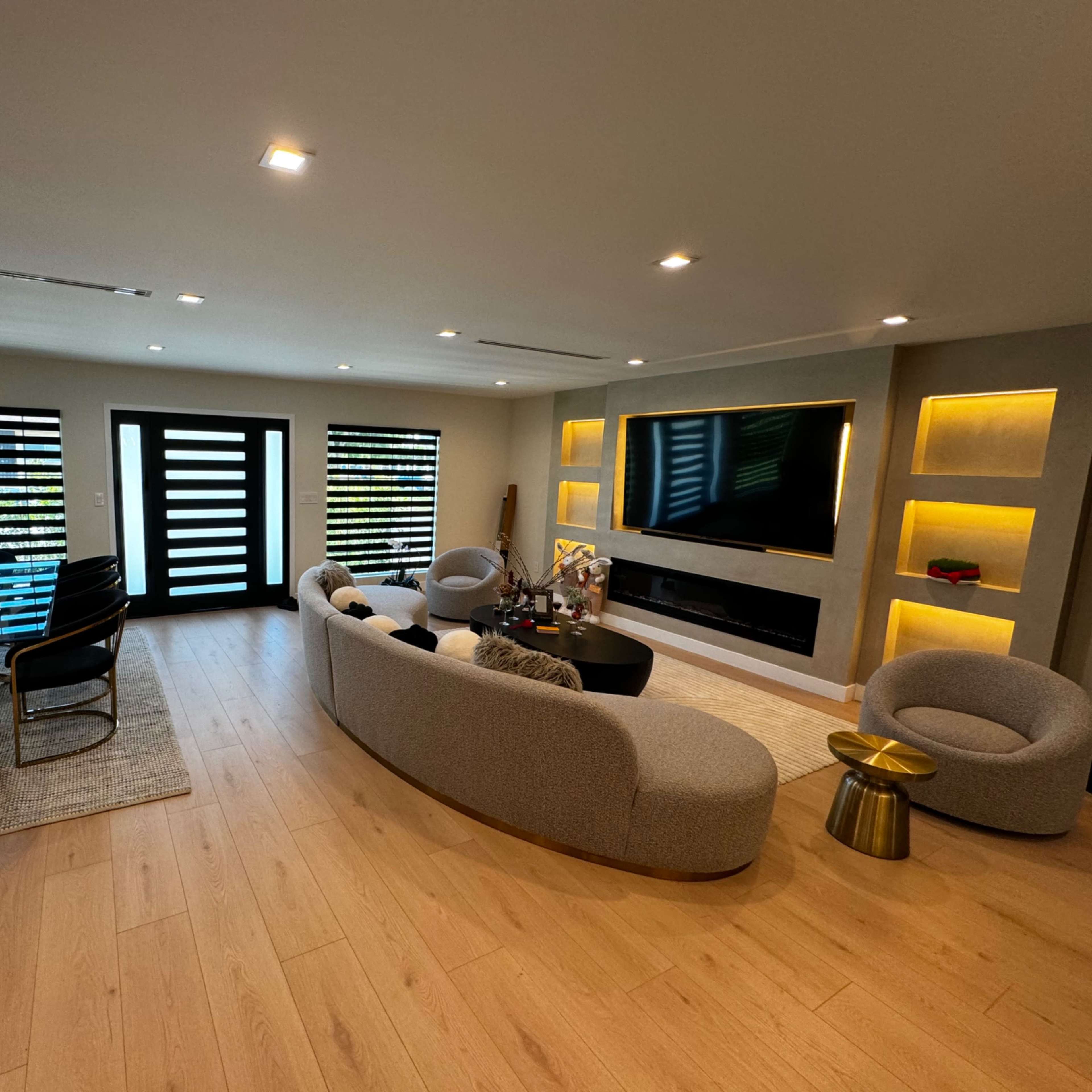 A modern living room with a curved sofa, accent chairs, a black coffee table, and a dining area featuring a glass table and black chairs, illuminated by built-in lighting.
