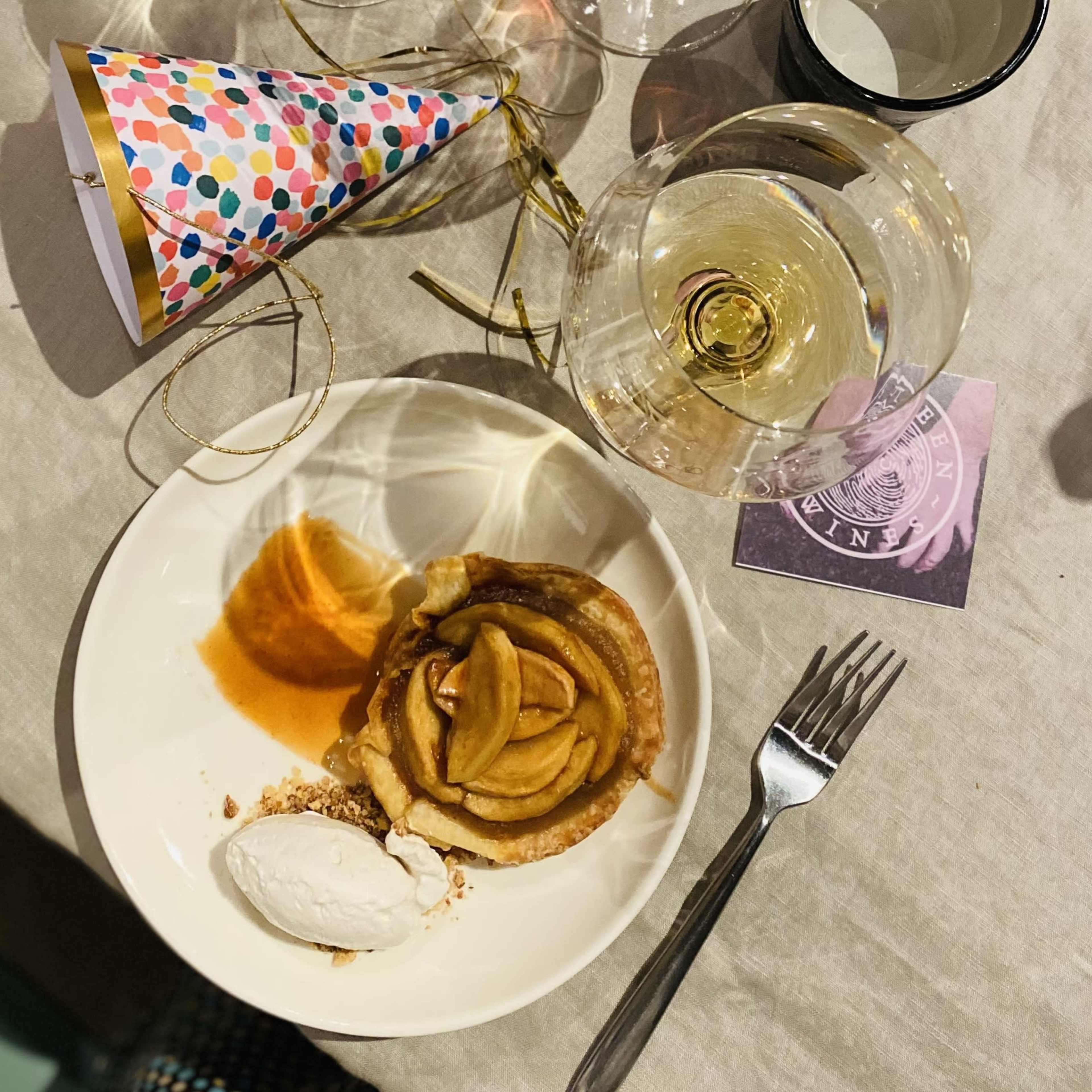 A plate with a slice of warm apple tart topped with a scoop of ice cream sits beside a wine glass and a colorful party hat on a table.