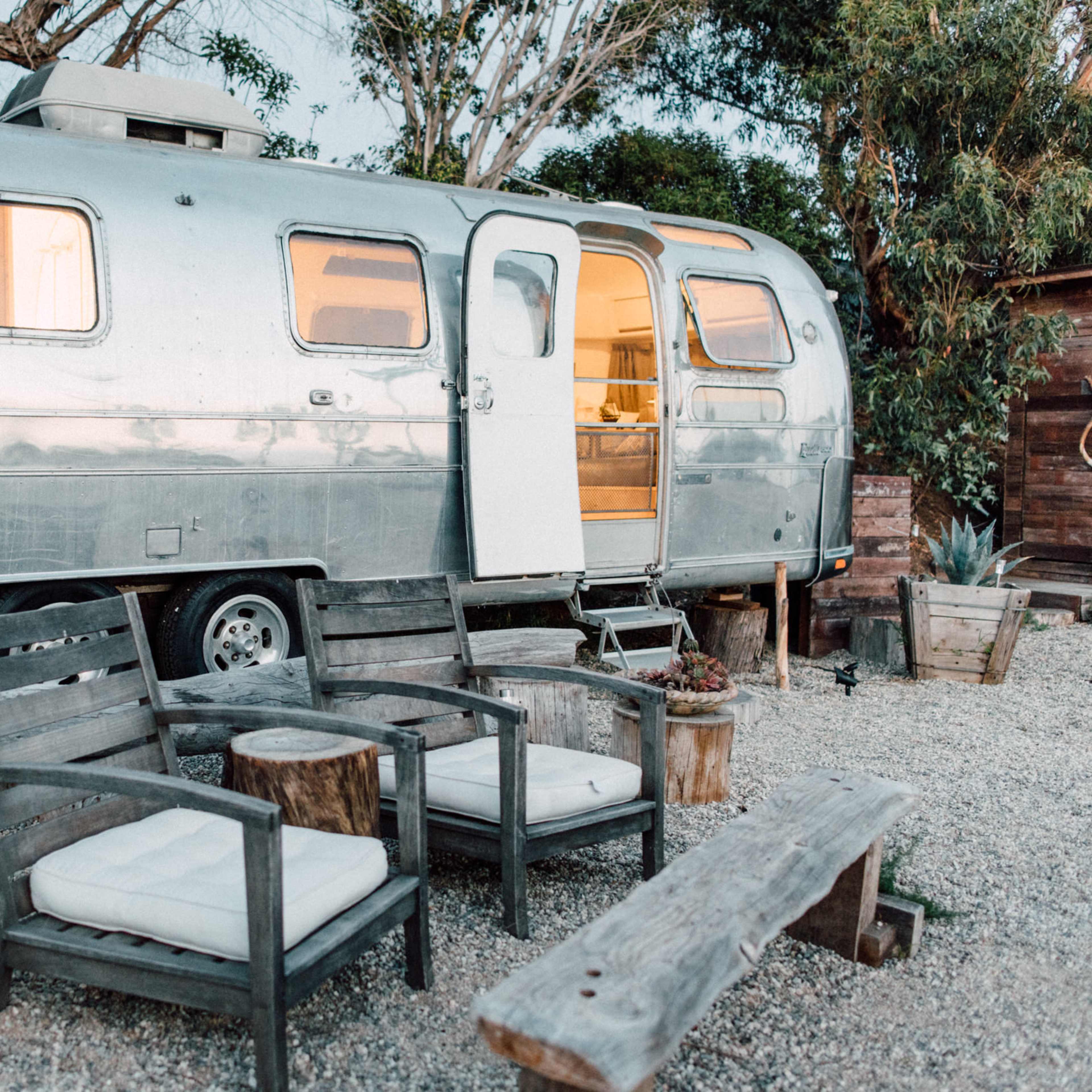 A silver Airstream trailer is parked beside rustic wooden chairs and tables, surrounded by plants and gravel.