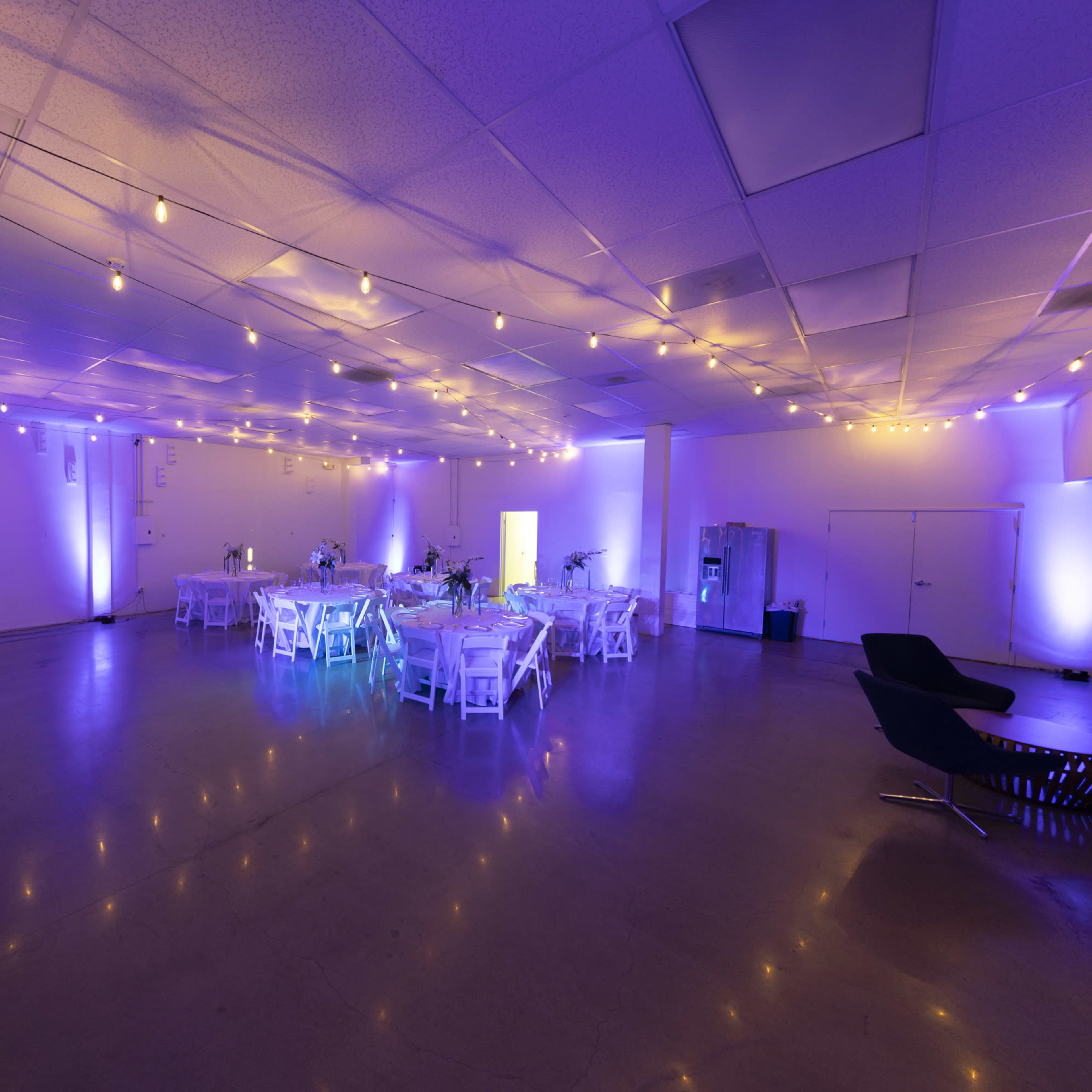 A spacious event venue features round tables with white tablecloths, decorated with centerpieces, and is illuminated by soft purple and warm white lighting, with string lights overhead.