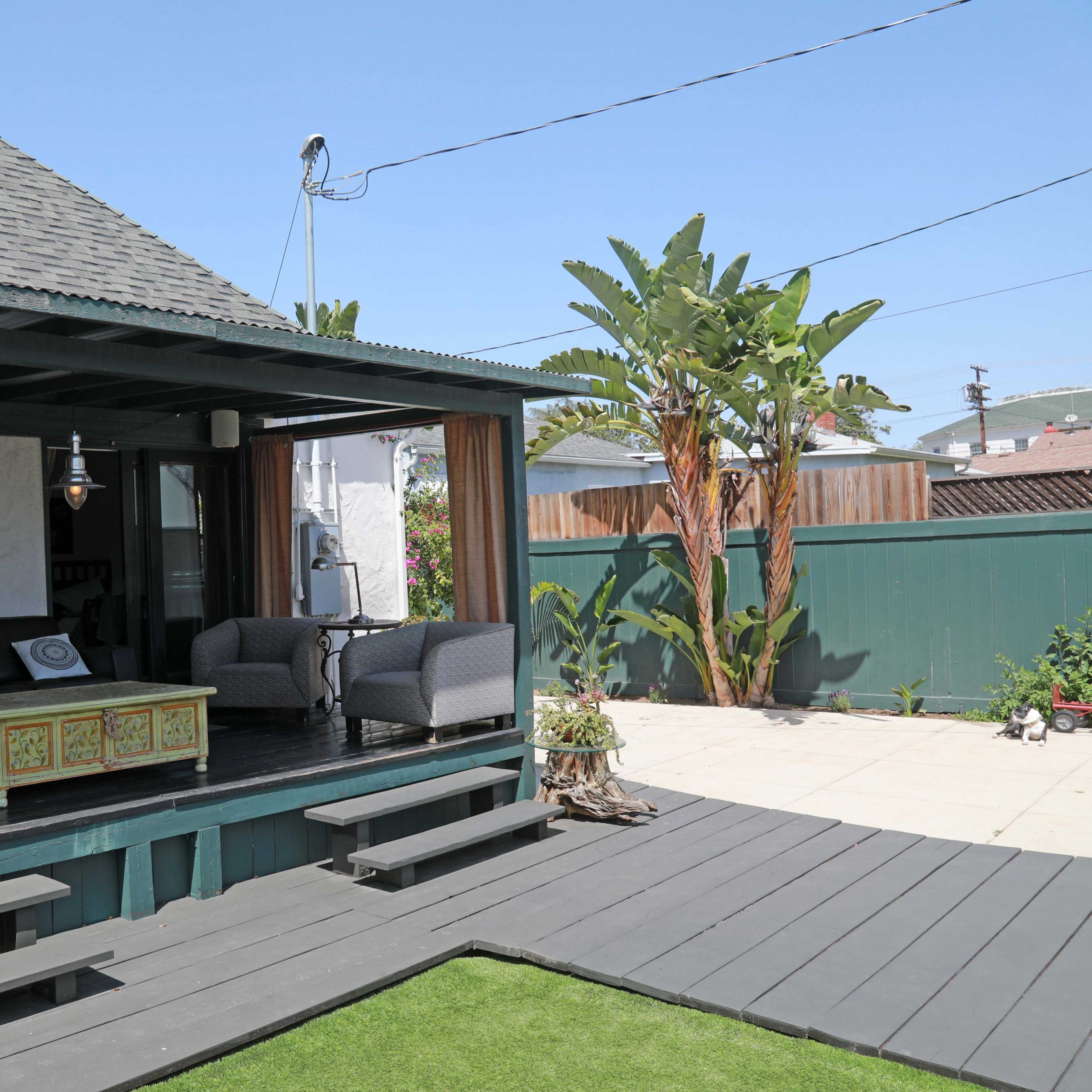 A shaded outdoor deck area with a seating arrangement and tropical plants in a residential backyard.