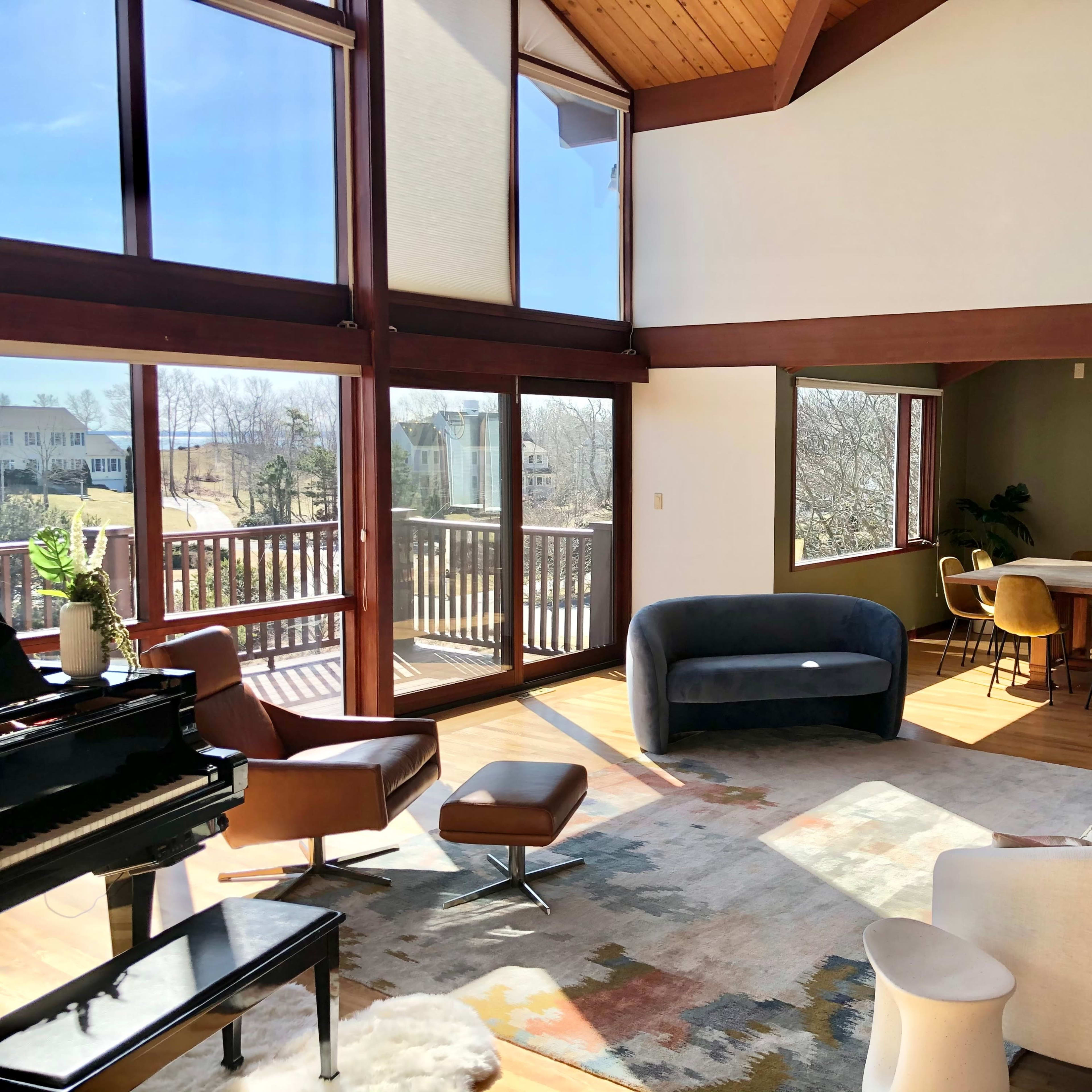 A modern living space featuring large windows, a piano, and various seating options, with a dining area visible in the background.