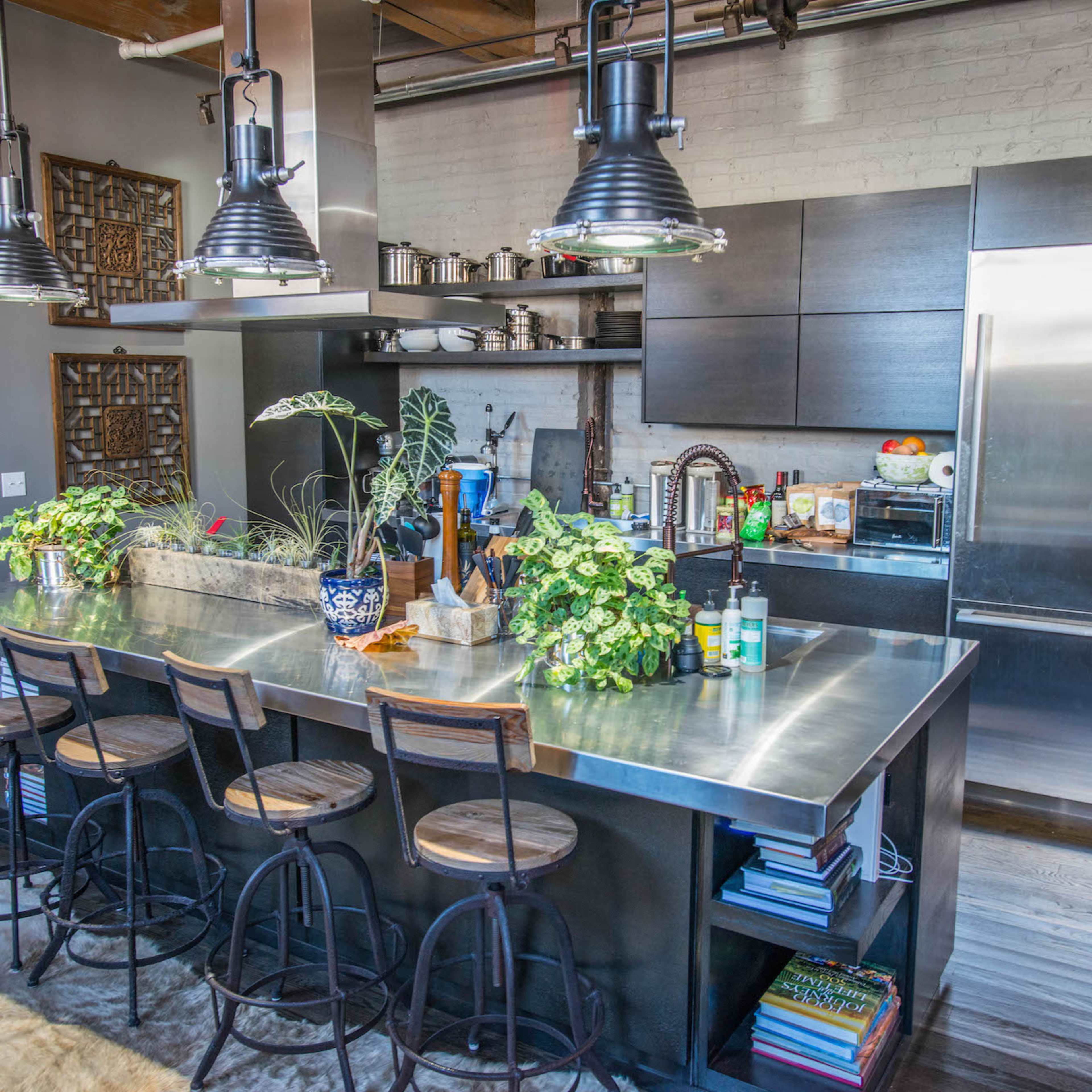 A modern kitchen with a central metal island, bar stools, and various plants, set against a backdrop of dark cabinetry and stainless steel appliances.