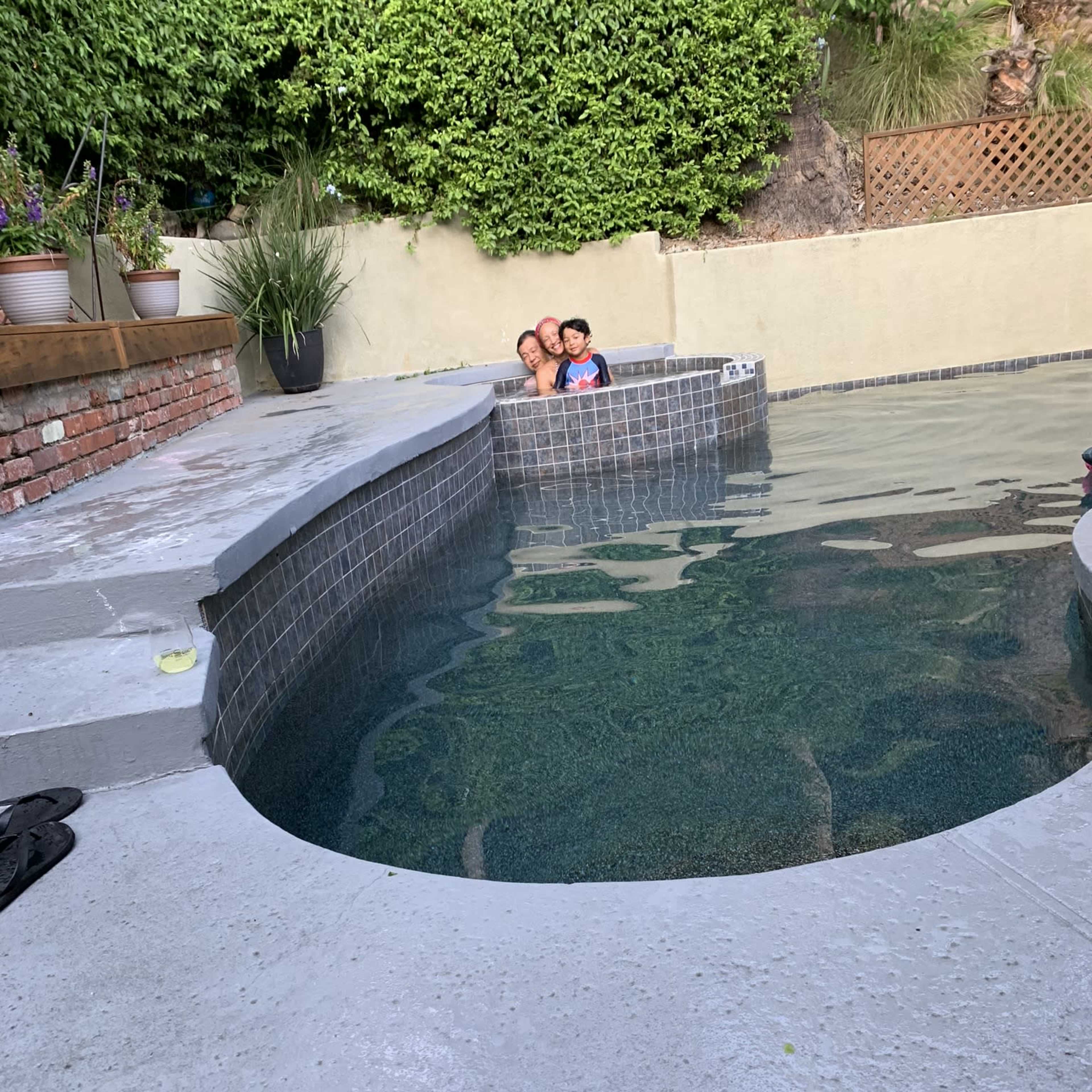 A child and an adult sit together on the edge of a hot tub beside a pool surrounded by greenery and a stone wall.