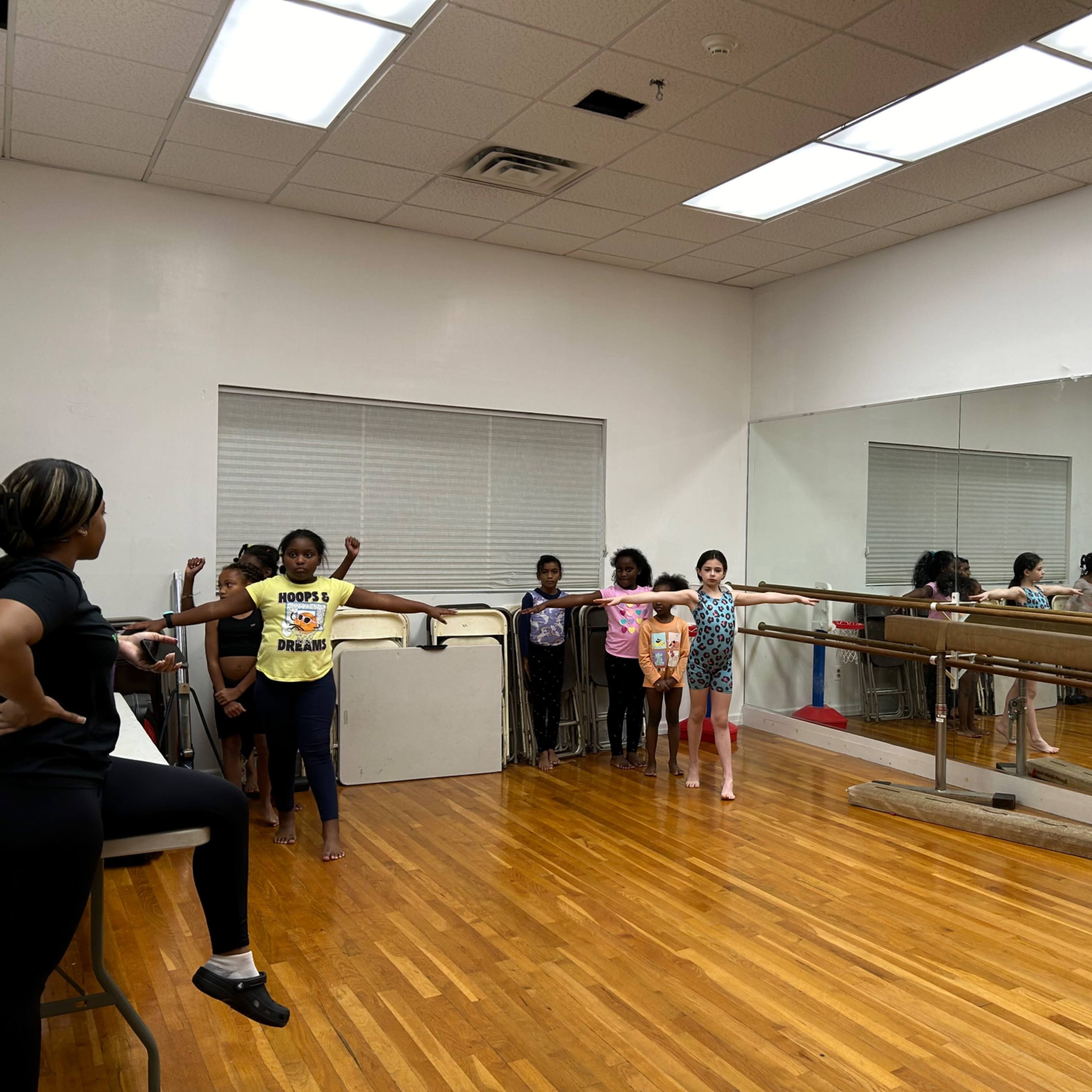Several children stand at a ballet barre in a dance studio with mirrors on the wall, while an instructor looks on.