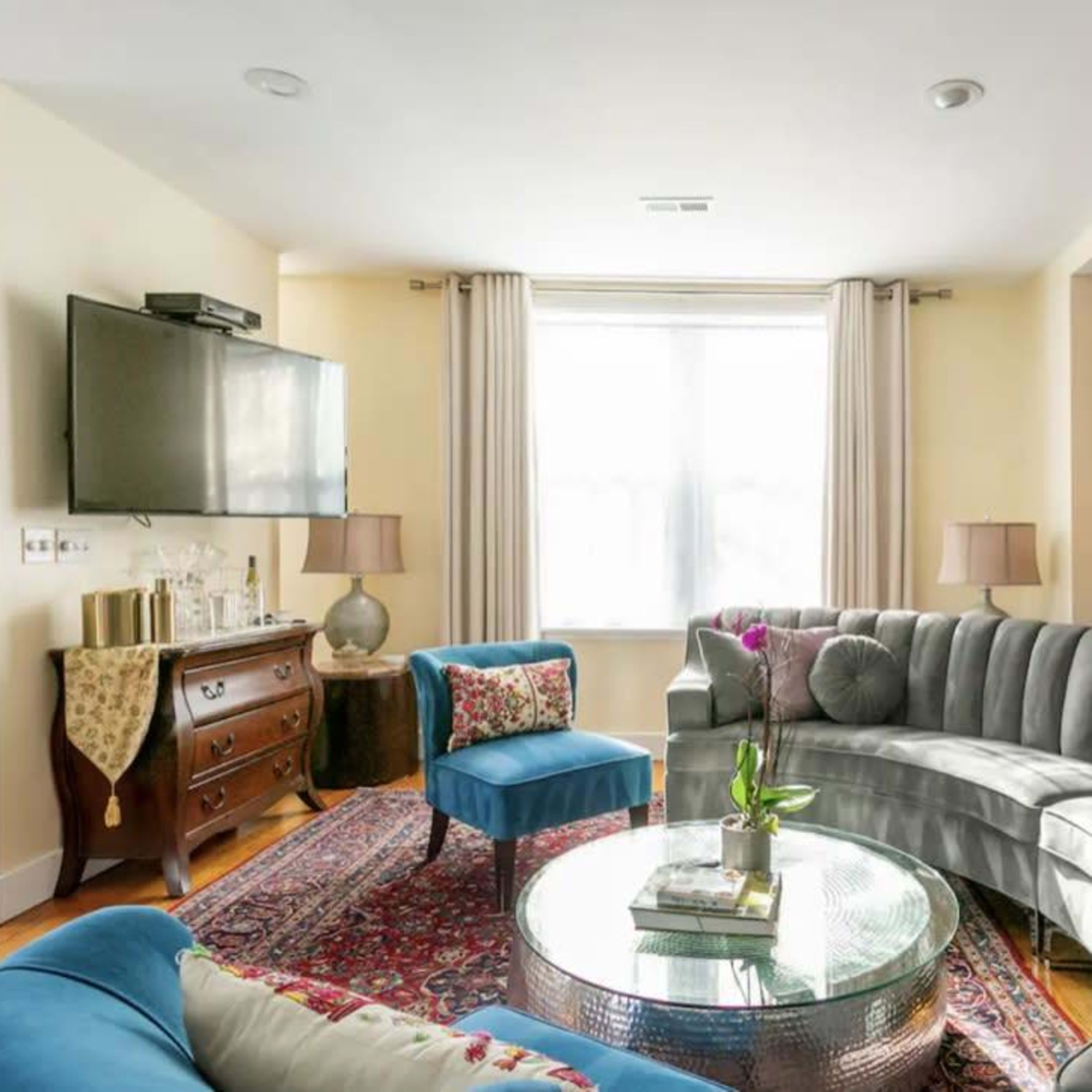 The living room features a curved gray sofa, a round glass coffee table, and a combination of colorful seating, with a window illuminating the space.