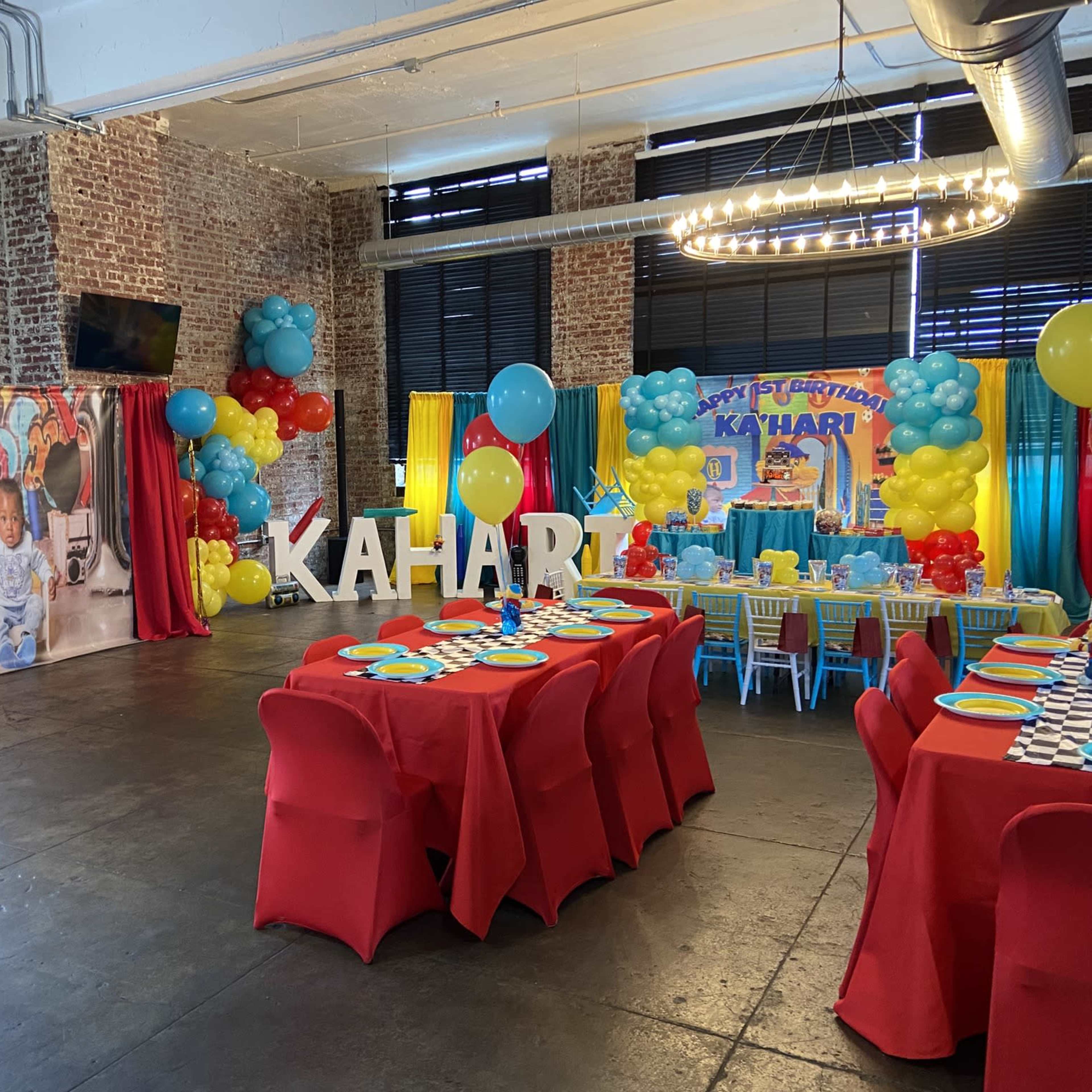 A vibrant birthday party setting with red and blue decorations, tables set with colorful tableware, and a backdrop featuring balloons and a large banner displaying the name "KAHARI."