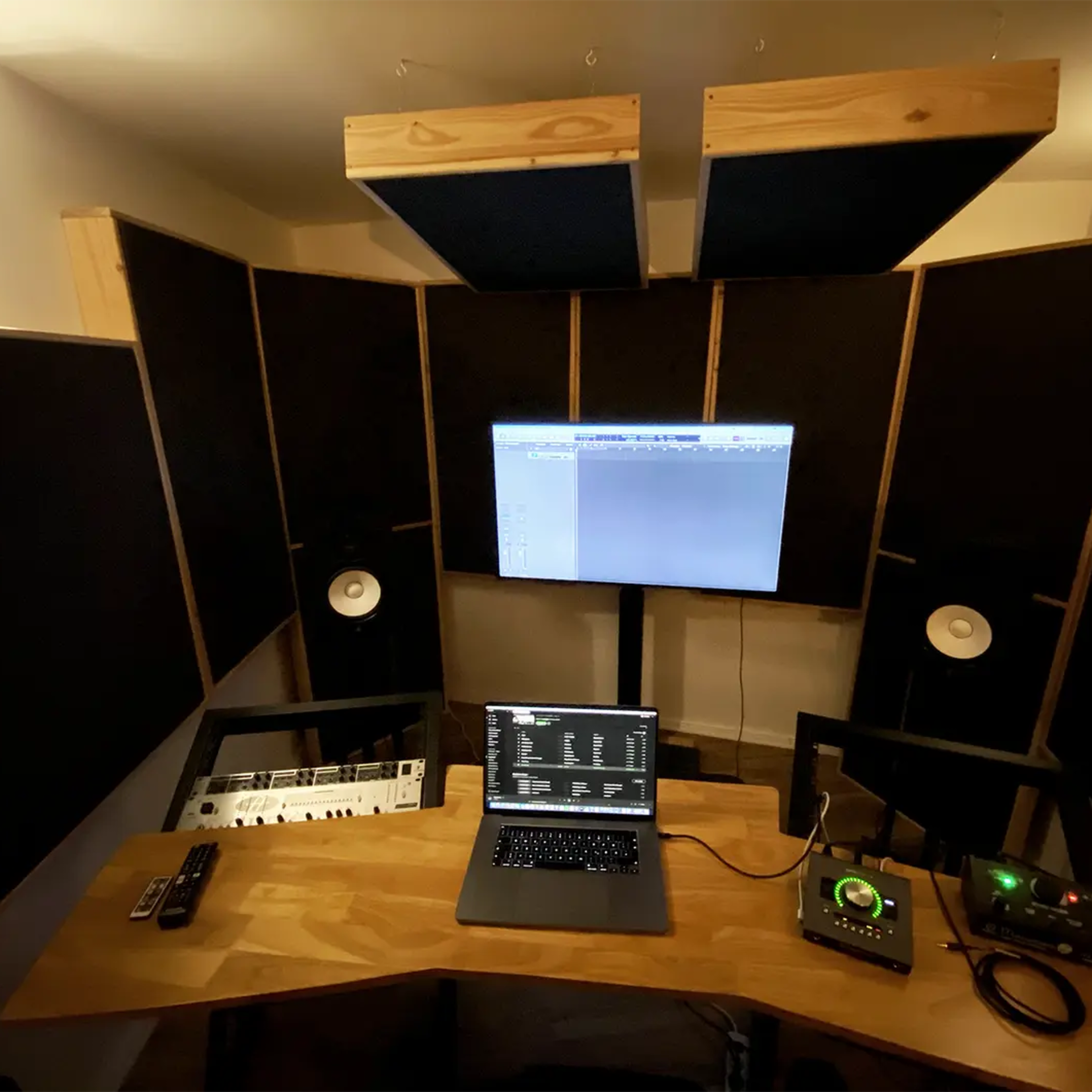 A music production studio is set up with a computer, audio interface, MIDI keyboard, and speakers arranged on a wooden desk surrounded by acoustic panels.