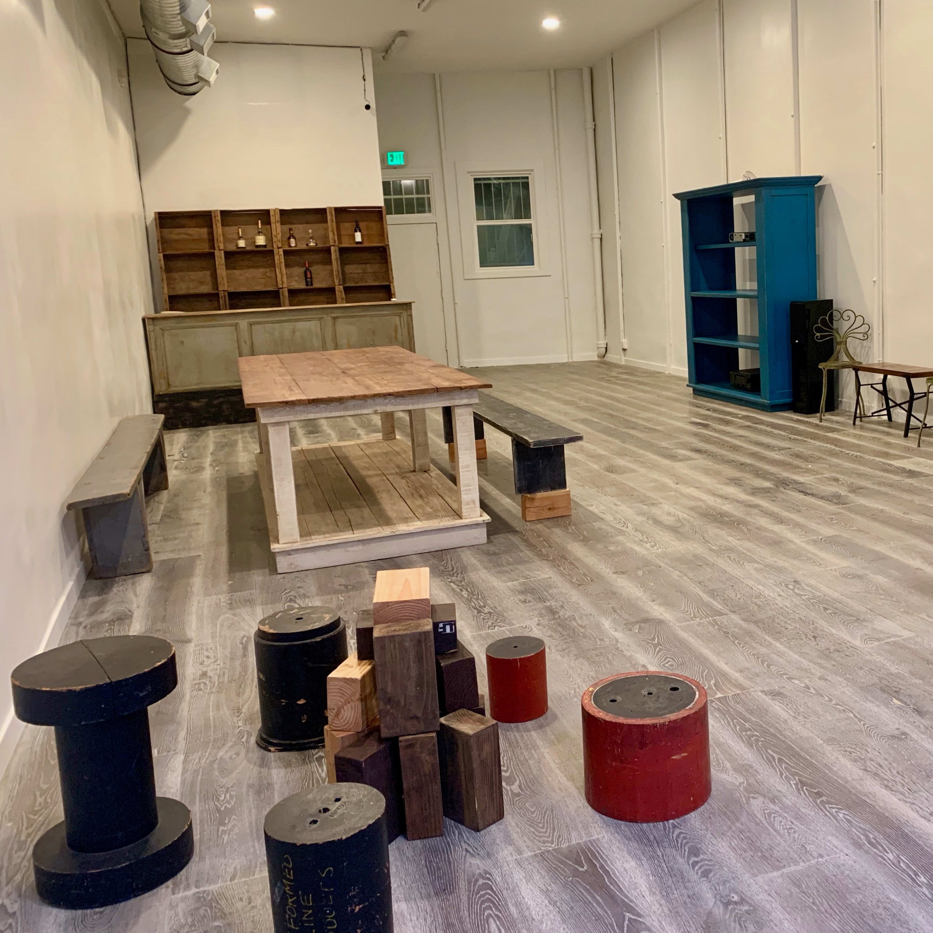 An empty room with wooden flooring, two blue bookcases, a wooden table, various cylindrical stools, and a set of shelves against the wall.