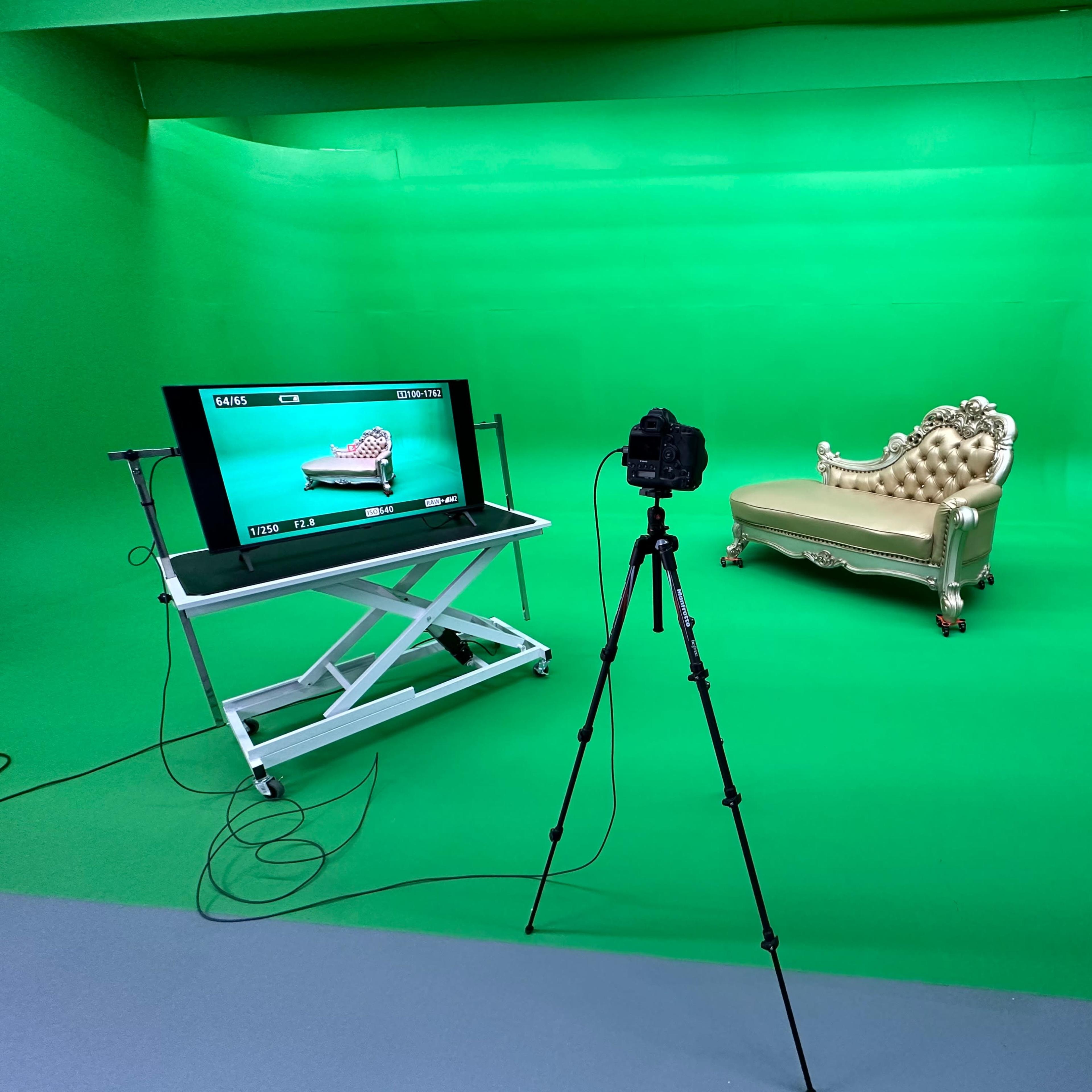 A photography setup in a green screen studio featuring a decorative chaise lounge, a camera on a tripod, and a monitor displaying a camera view.