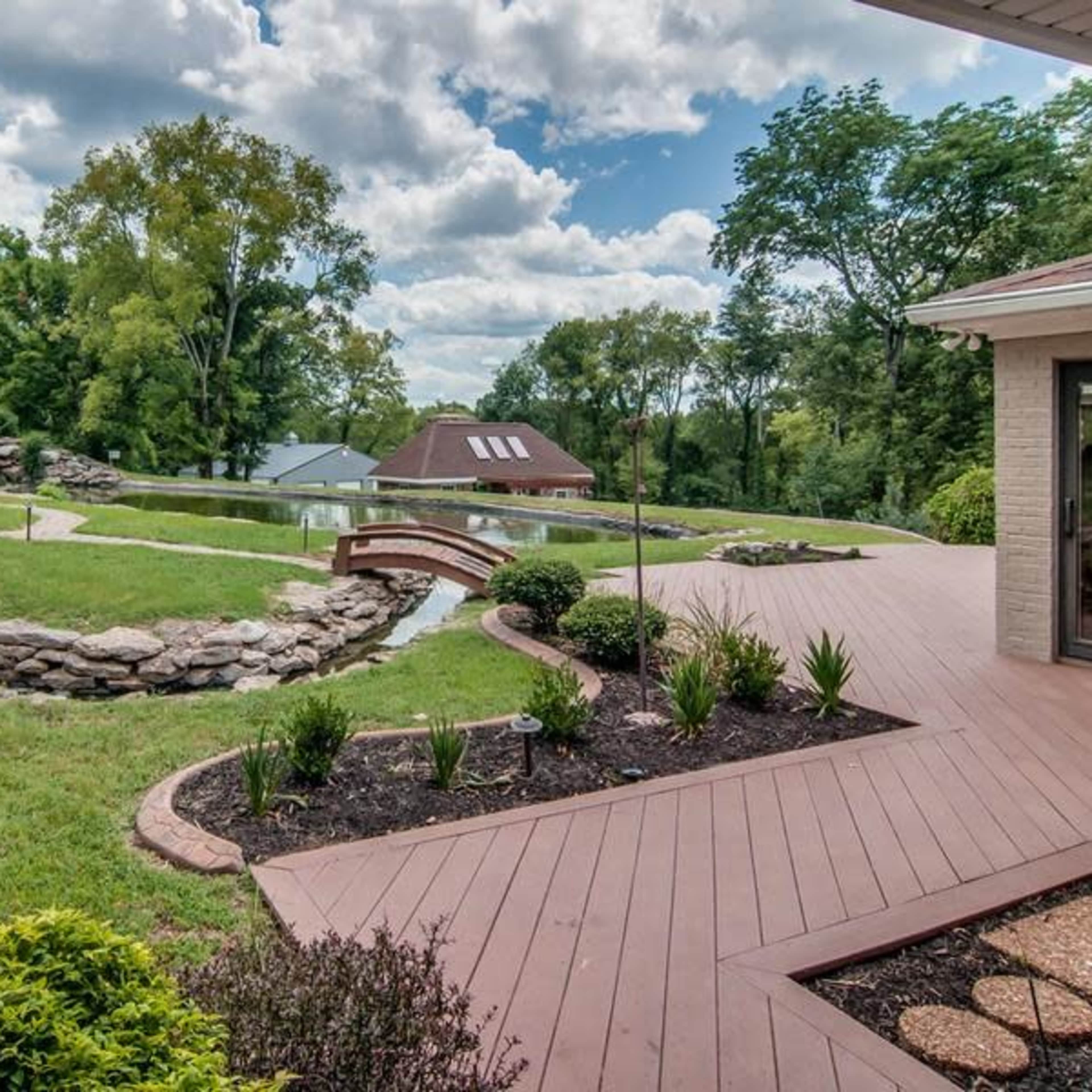 A landscaped backyard features a wooden deck winding around a stone-edged pond surrounded by greenery and trees.