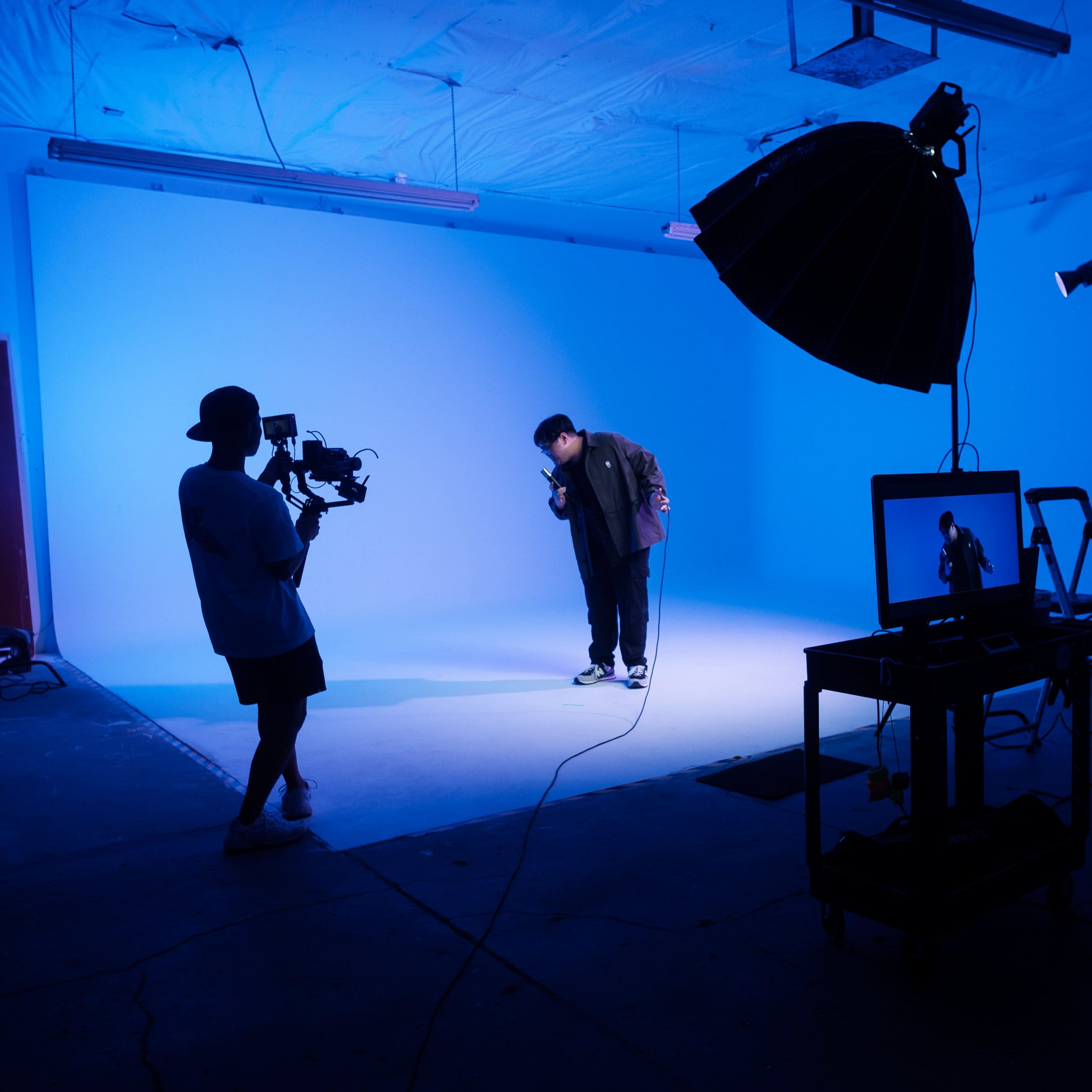 A person is performing on a white floor in a studio illuminated by blue lighting, while another individual films them using a camera on a tripod.