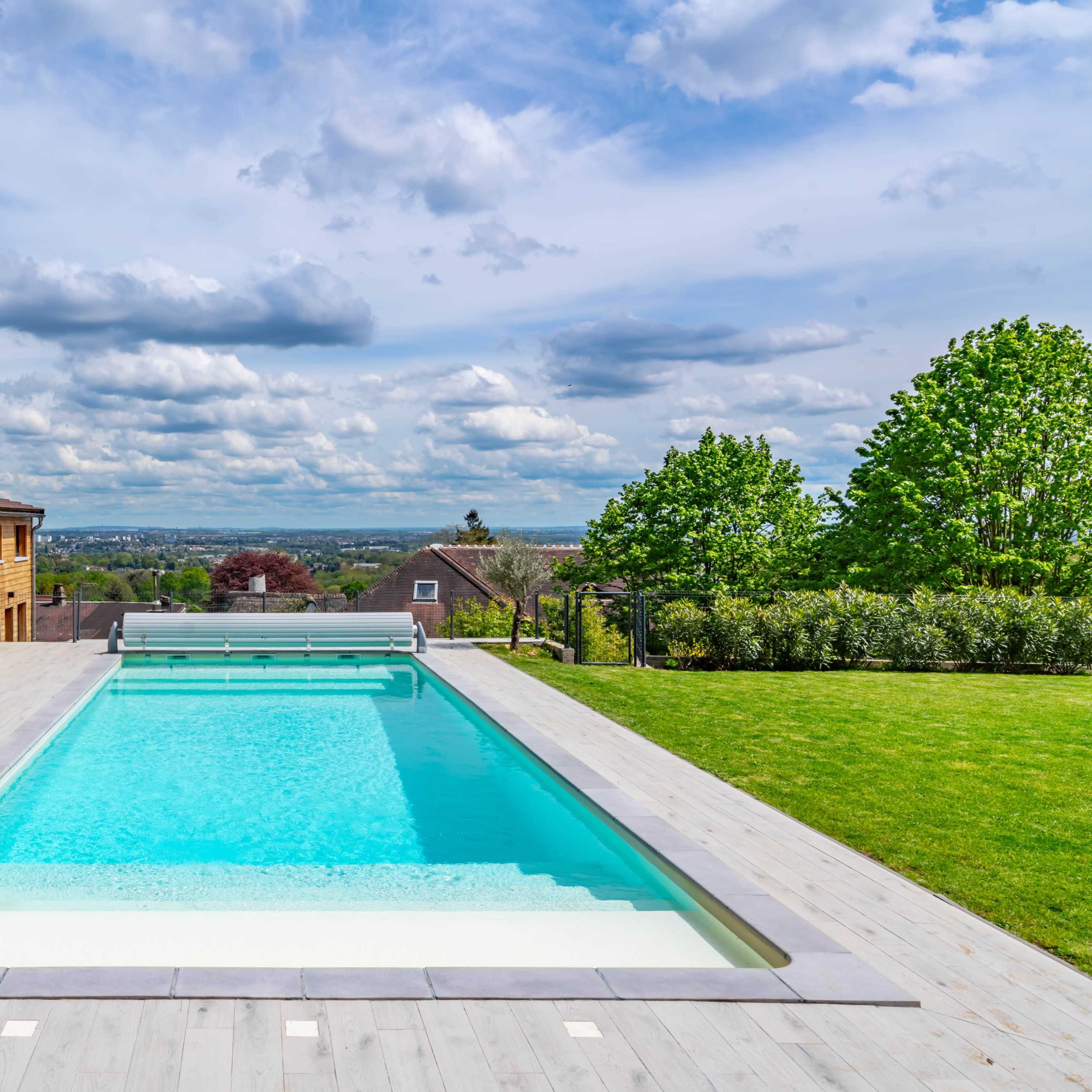 A modern swimming pool with a wooden deck is set against a backdrop of green trees and rolling hills under a partly cloudy sky.