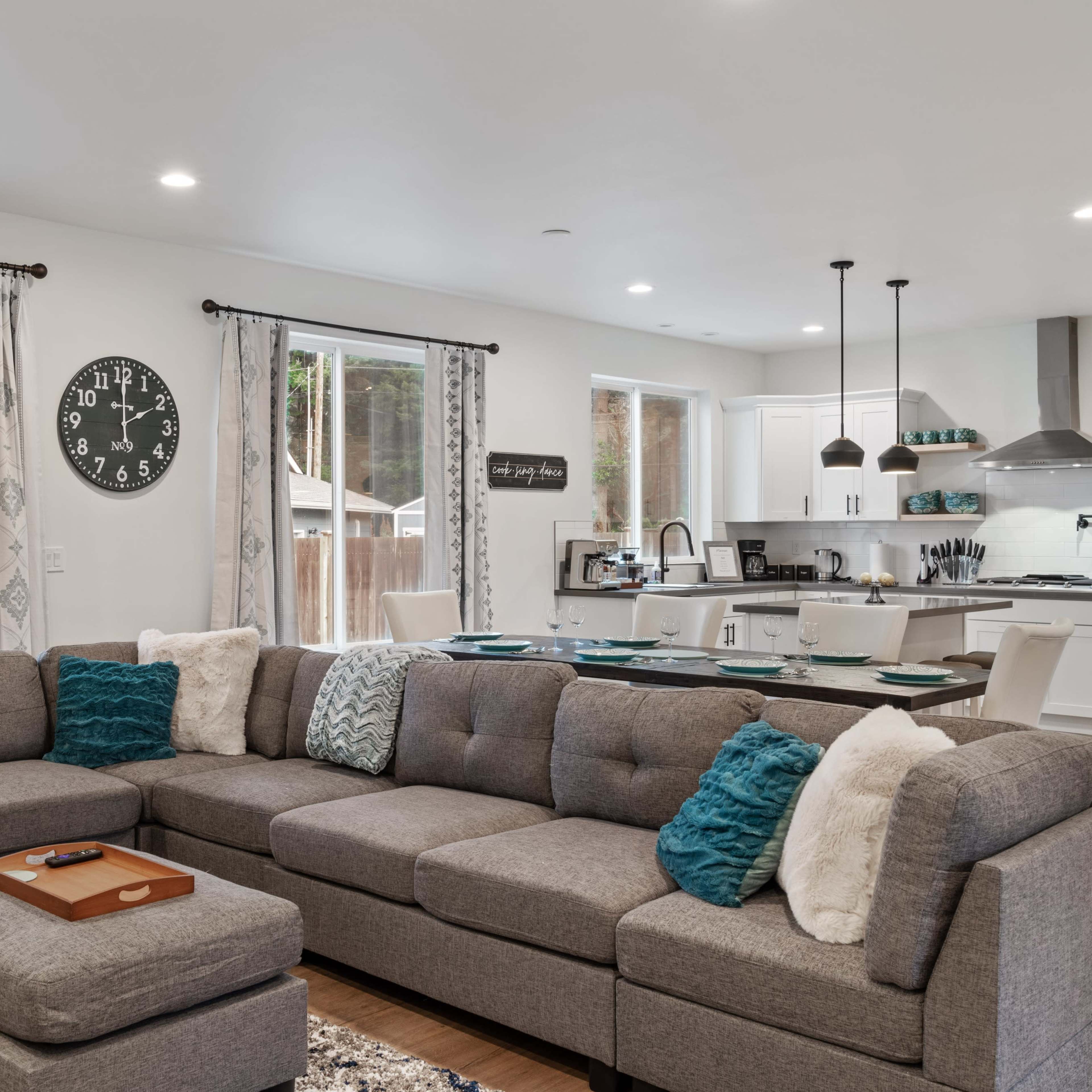 A modern living room features a large gray sectional sofa with decorative pillows, an ottoman, and an open layout connecting to a kitchen area.