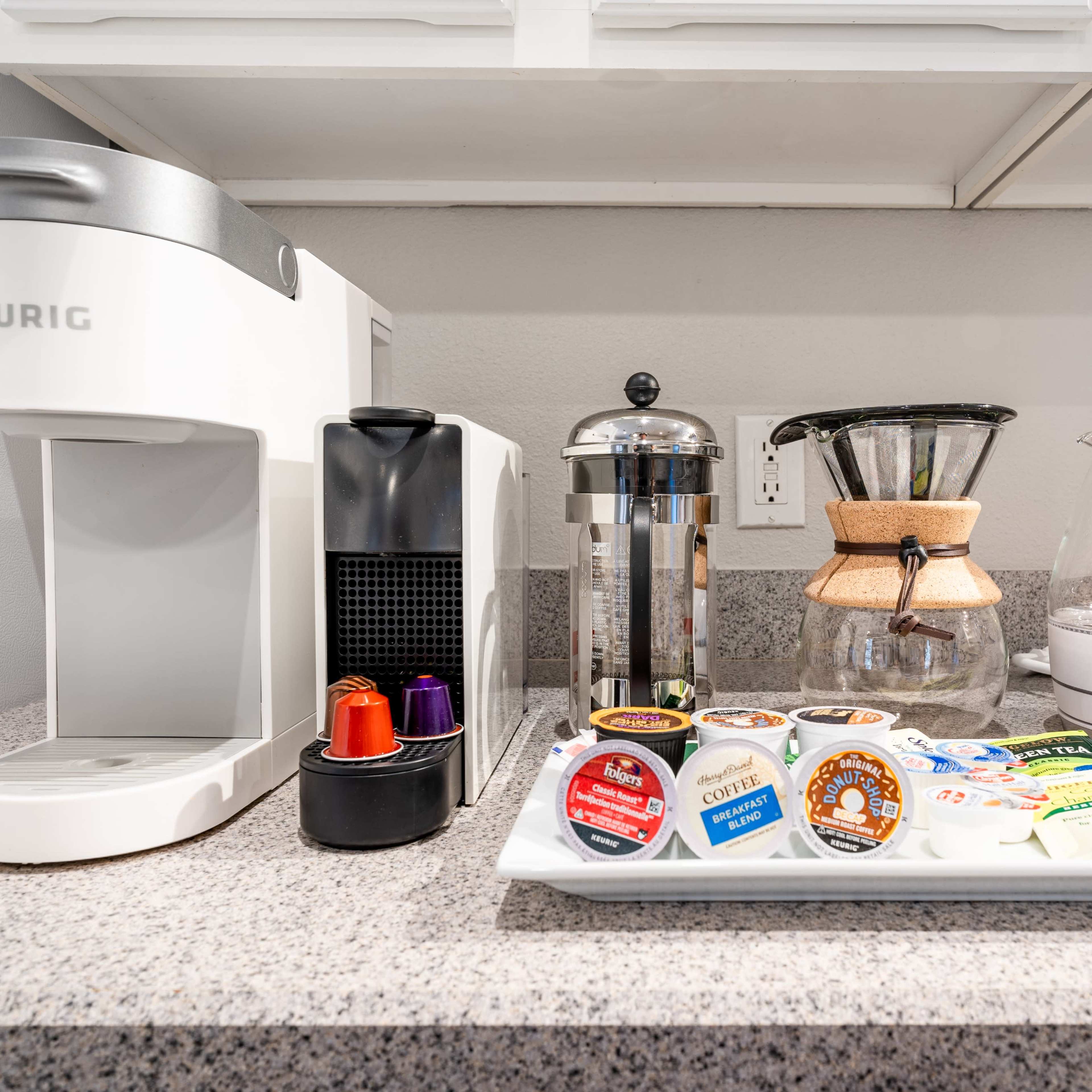 A countertop features a Keurig coffee maker, a French press, an electric kettle, and a tray with various coffee and tea pods.