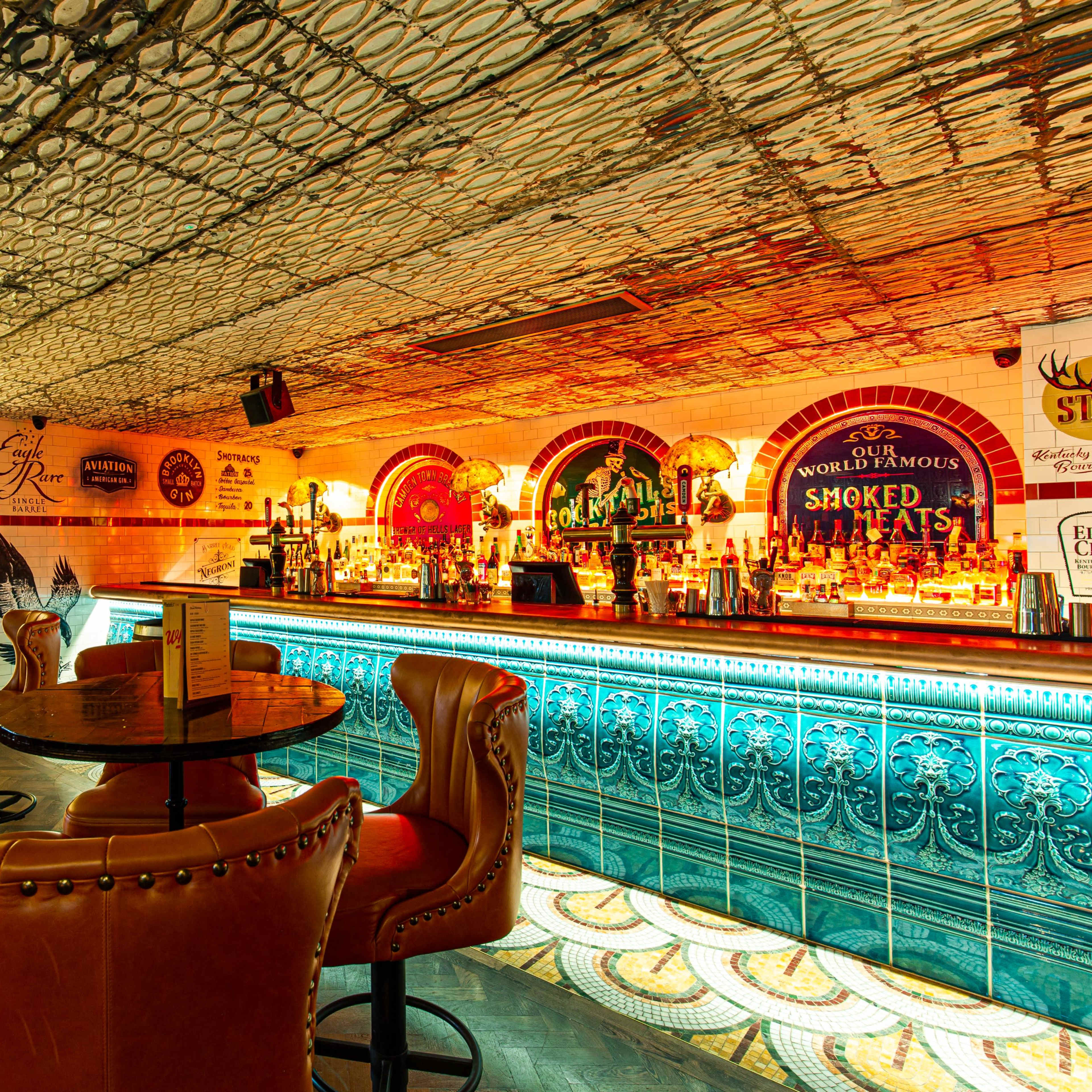 A stylish bar area with ornate tile work, high-backed leather chairs, and a countertop lined with various bottles and illuminated signage.